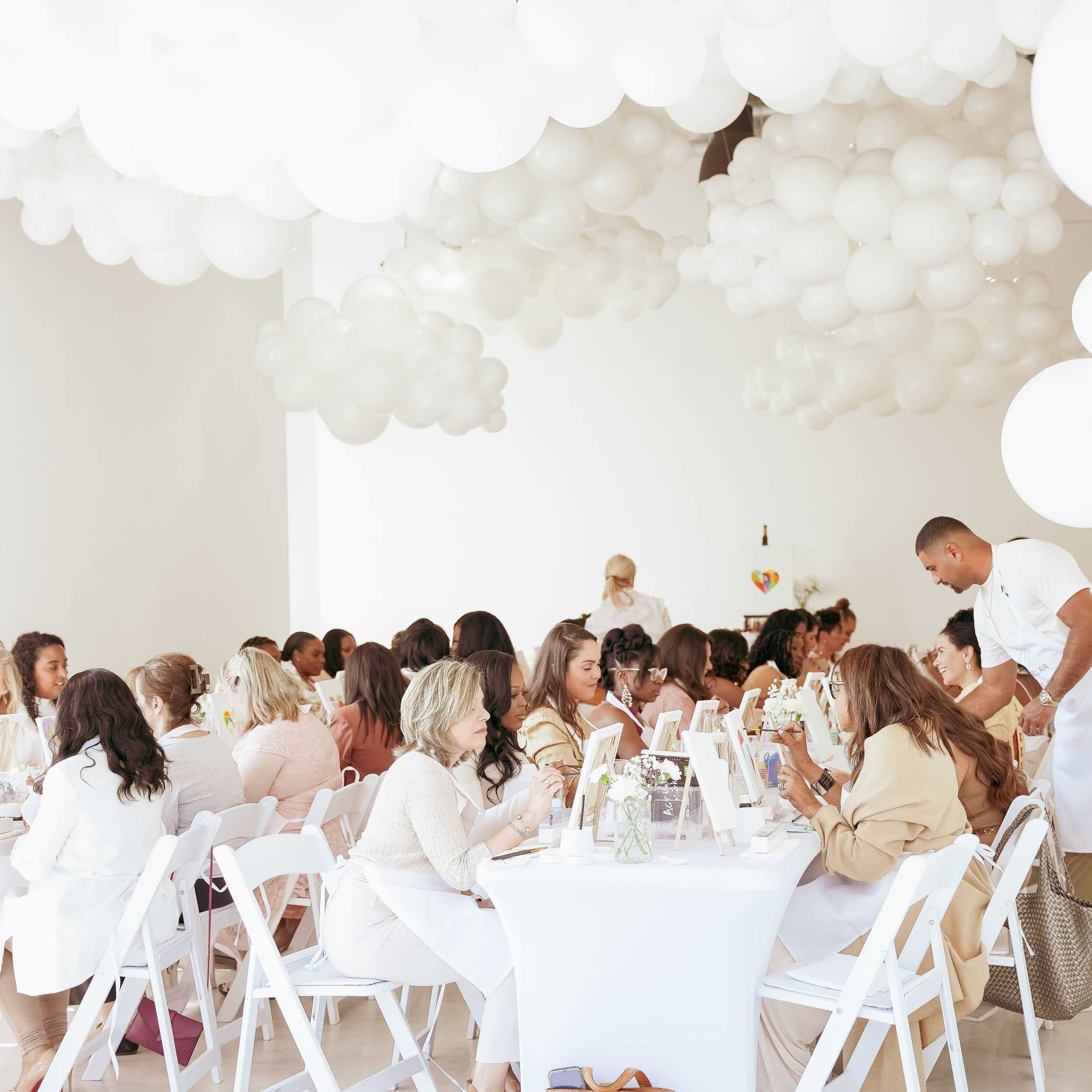 A gathering of women seated at long tables covered with white tablecloths in a room decorated with clusters of white balloons.