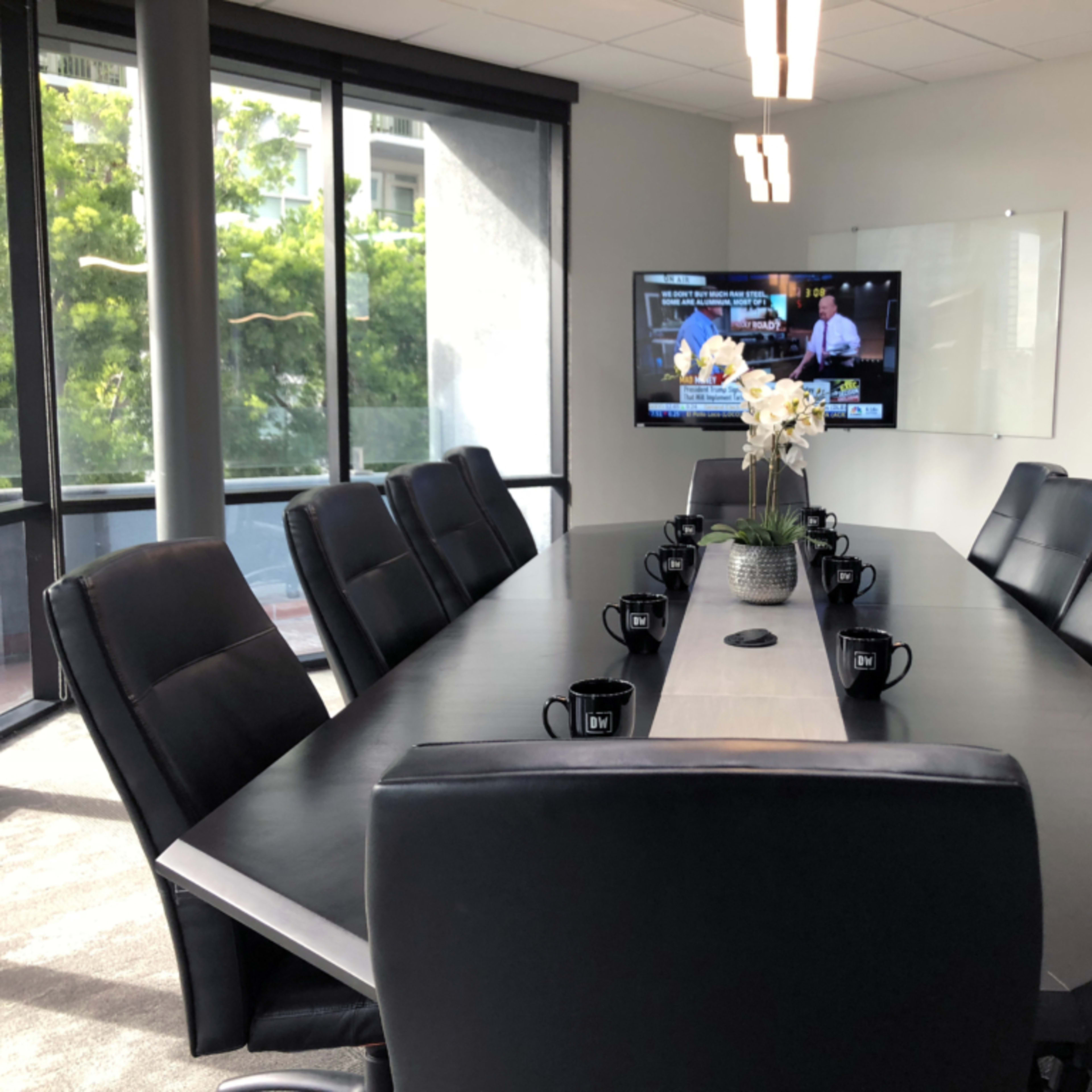 A modern conference room features a large oval table surrounded by black chairs, with a television displaying news in the background and a floral centerpiece on the table.