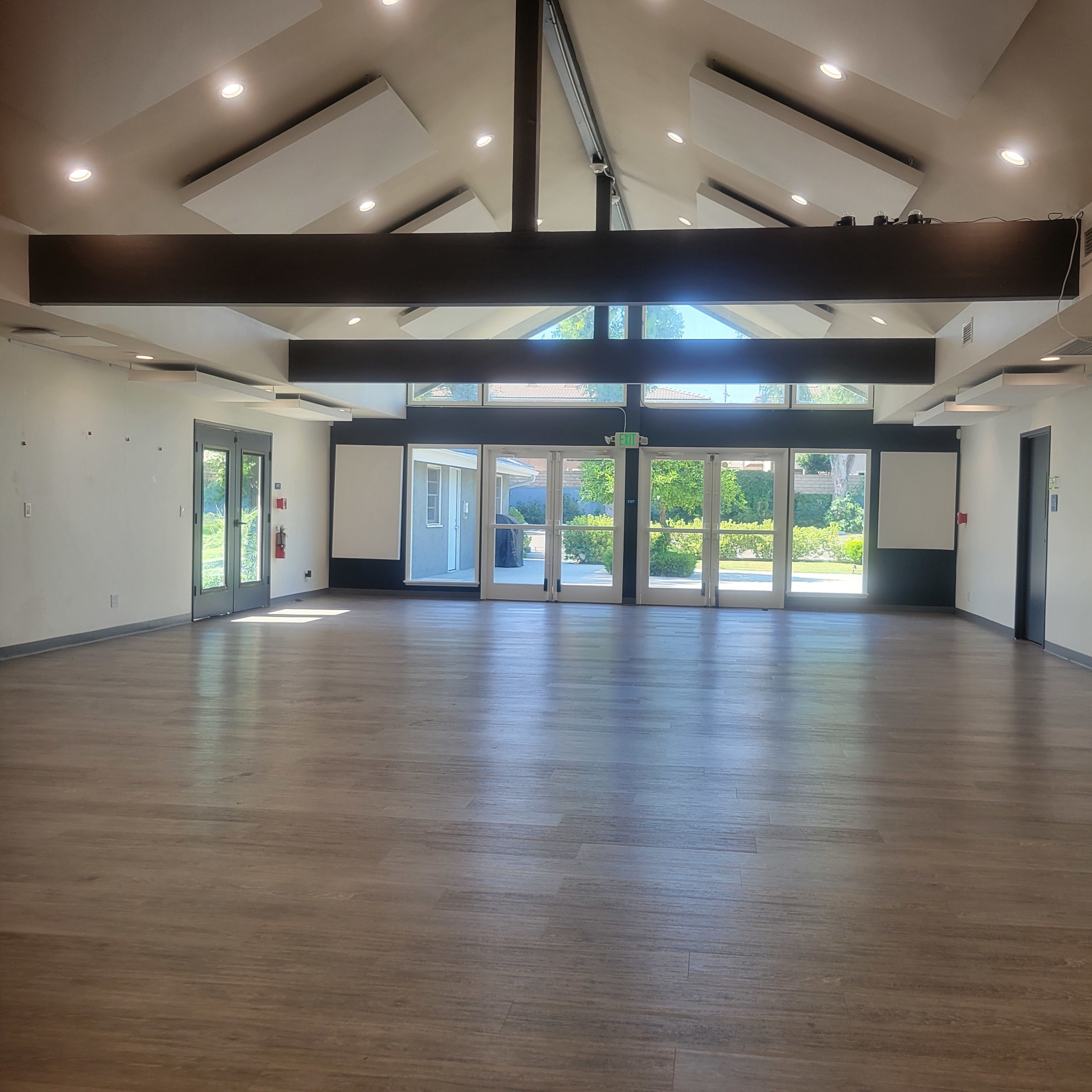 A large, empty indoor space with high ceilings, wooden flooring, and glass doors leading to an outdoor area.
