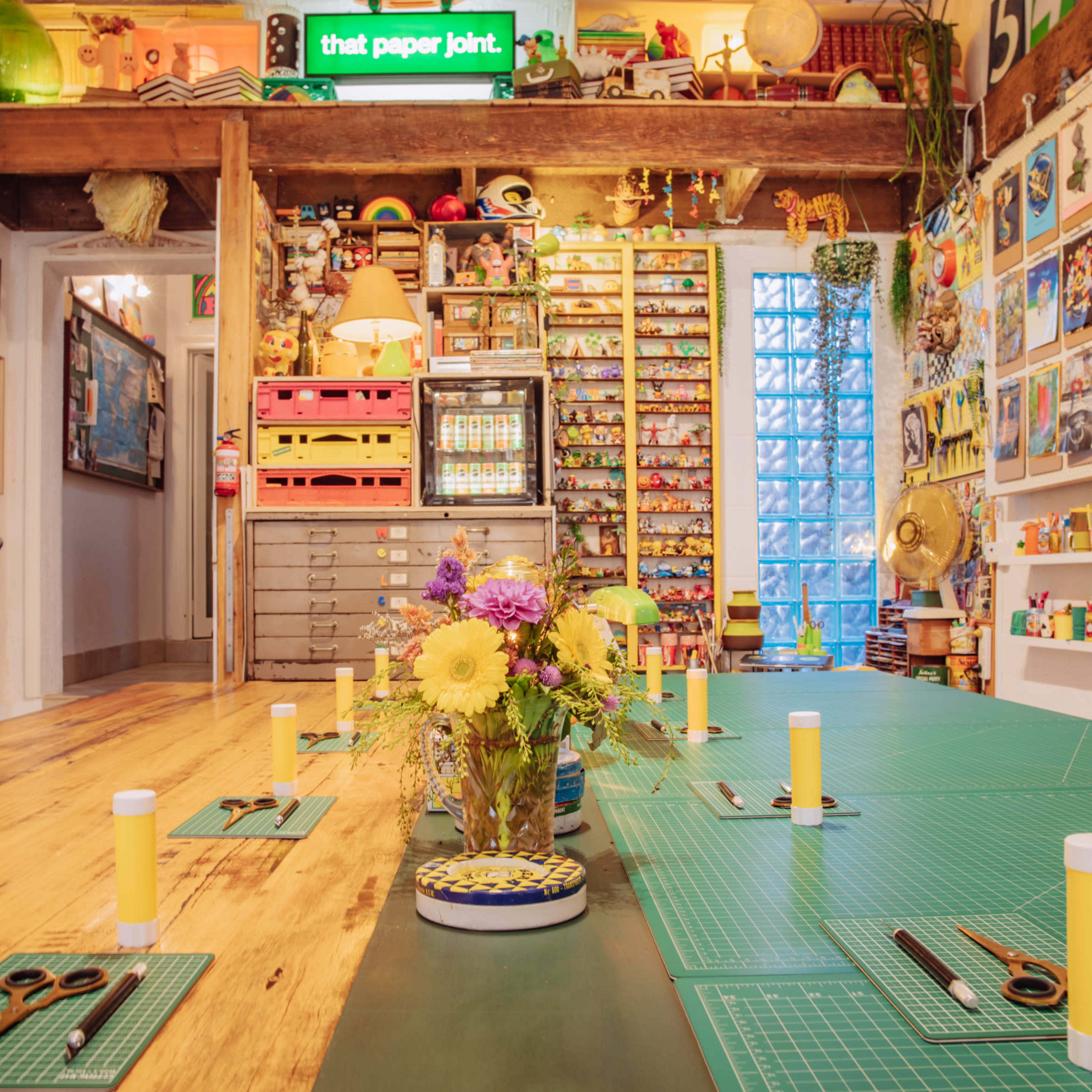 A colorful creative workspace filled with art supplies, a long table set for a project, and walls adorned with various framed artworks.