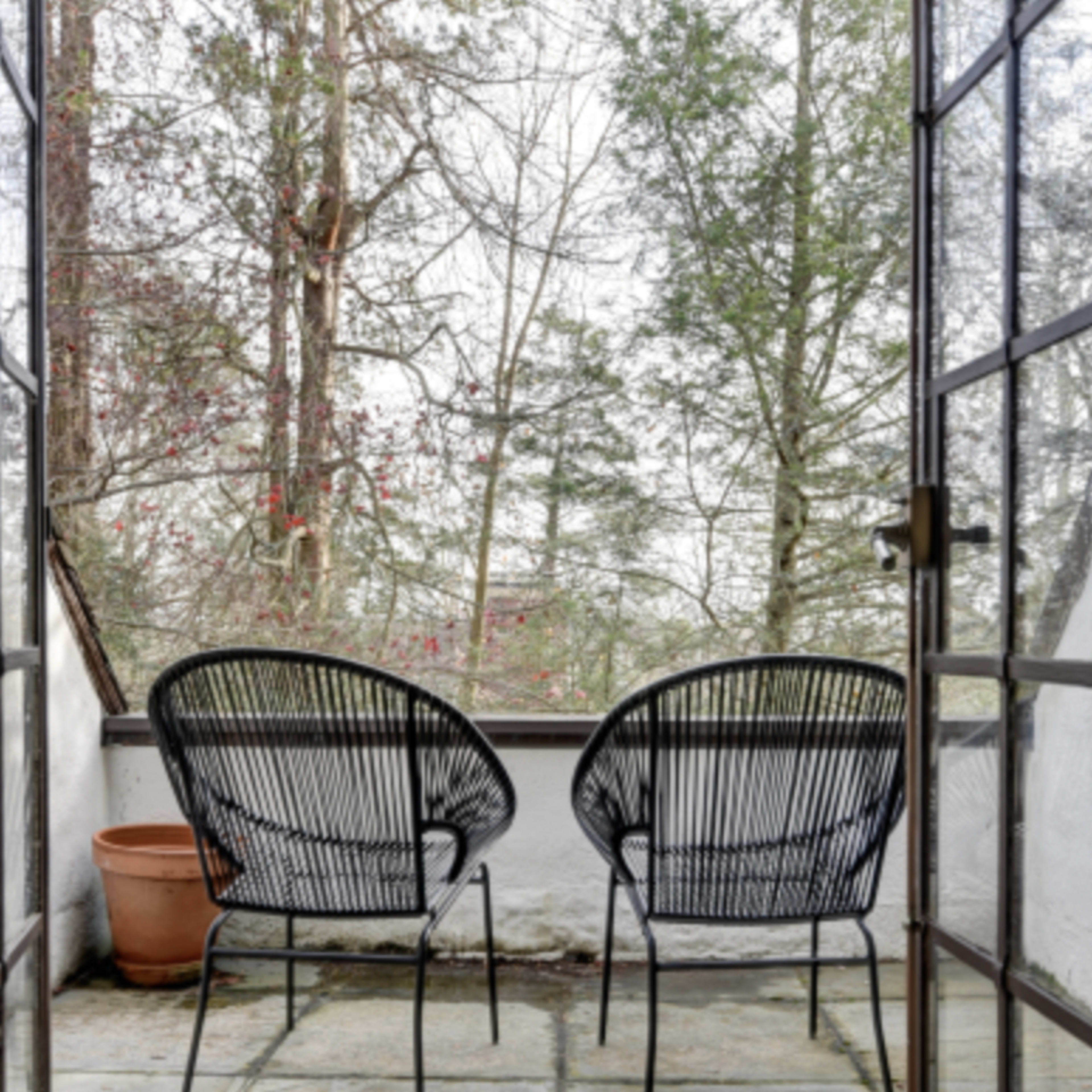 Two wicker chairs are positioned on a stone patio, with a view of a wooded area through a large open doorway.