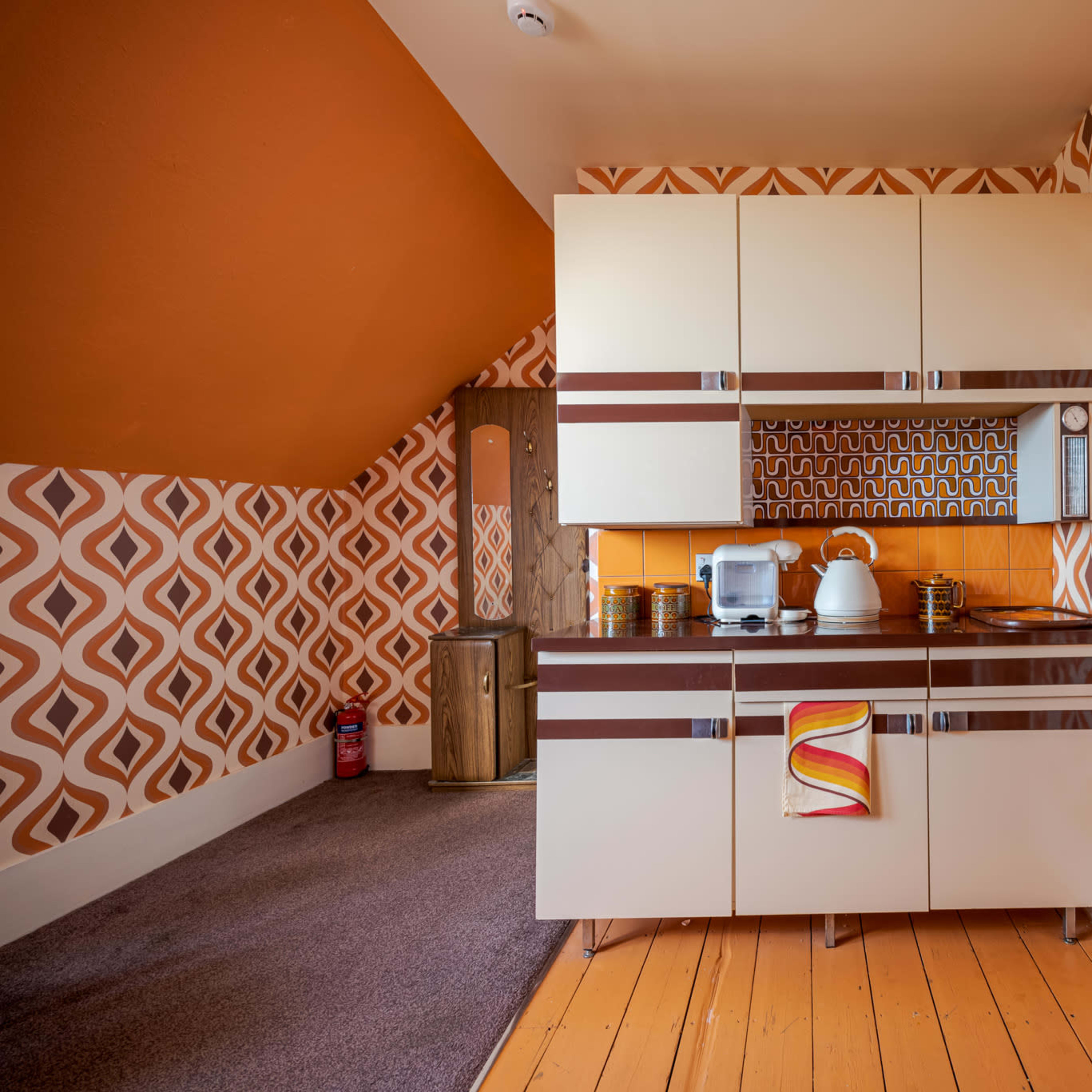 The kitchen features retro-style cabinetry with a patterned wallpaper in orange and brown tones, accompanied by wooden flooring.