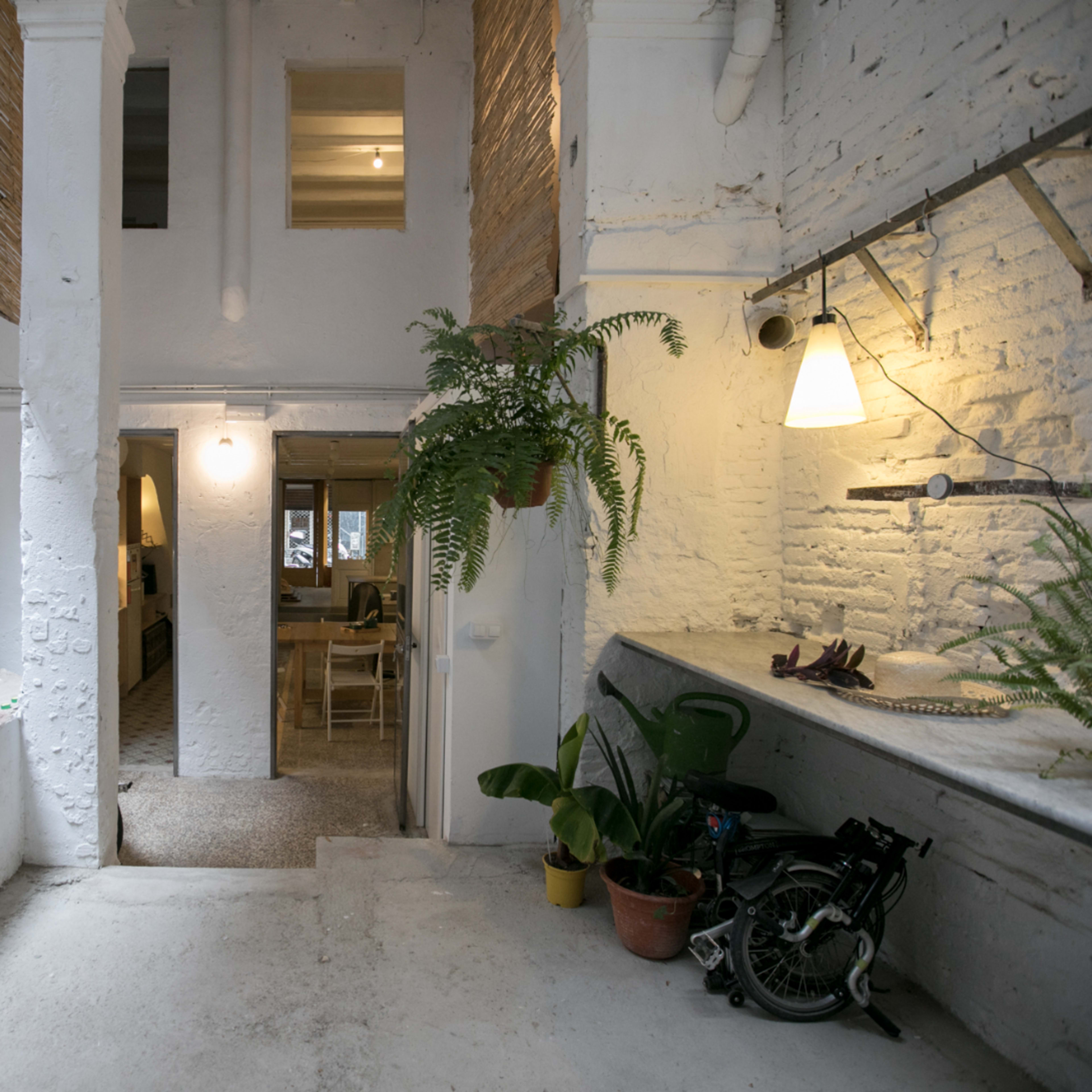 A spacious, white-walled interior featuring a small kitchenette on the right, a seating area visible in the background, and greenery accentuating the space.