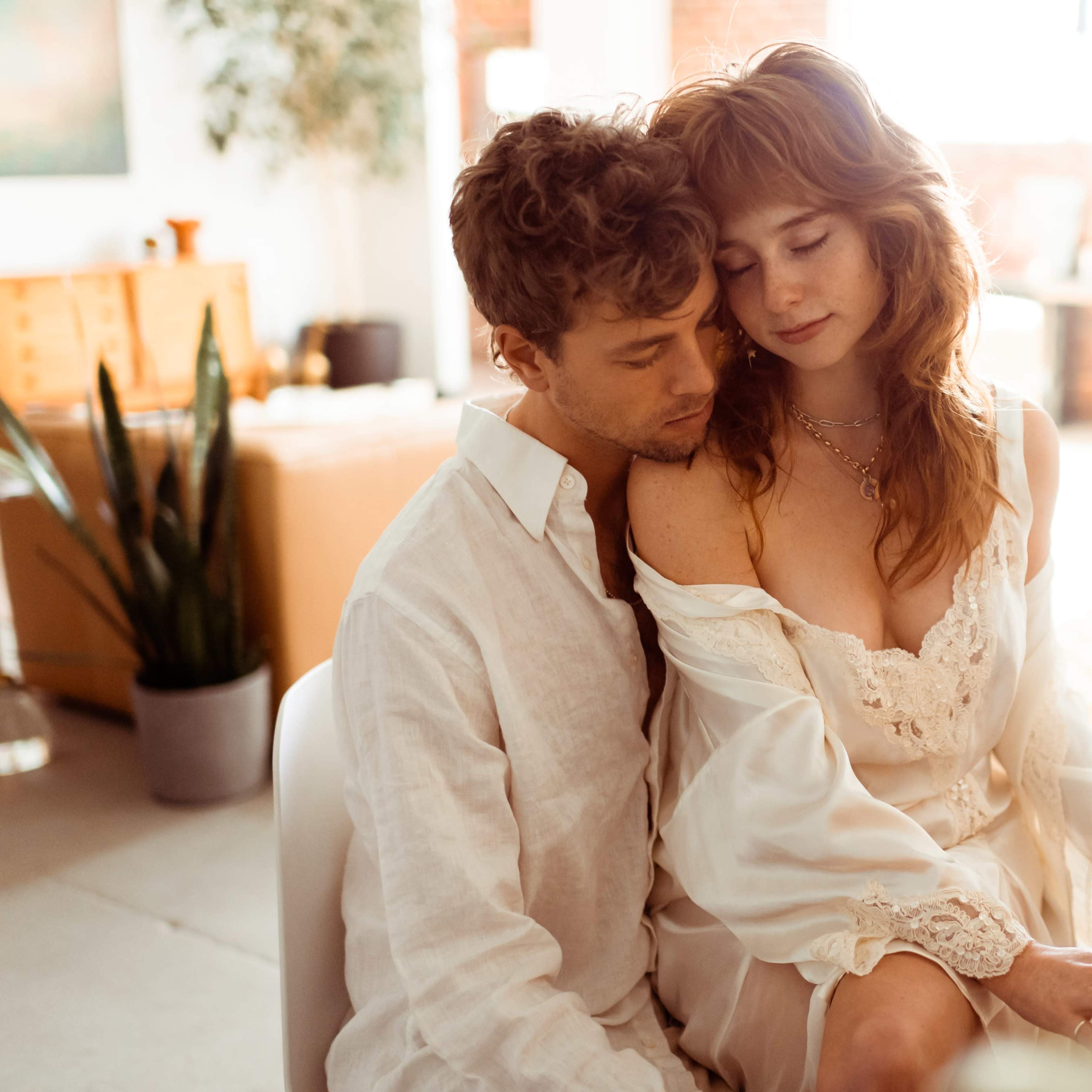 A couple sits together at a piano in a brightly lit, modern room with large windows and plants around them.