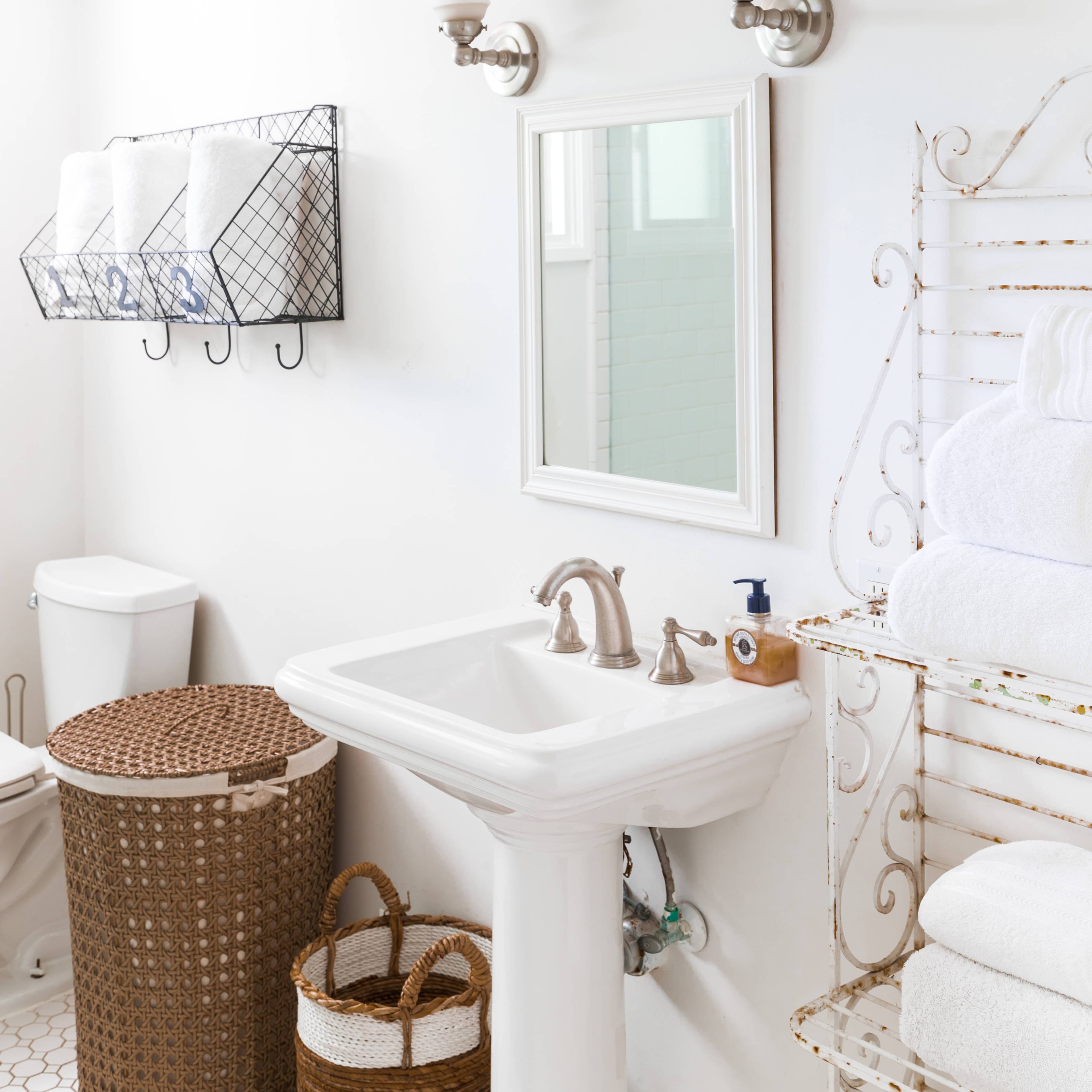A clean bathroom features a white sink, a mirror, neatly stacked towels, a toilet, and a basket for storage on a bright, uncluttered wall.