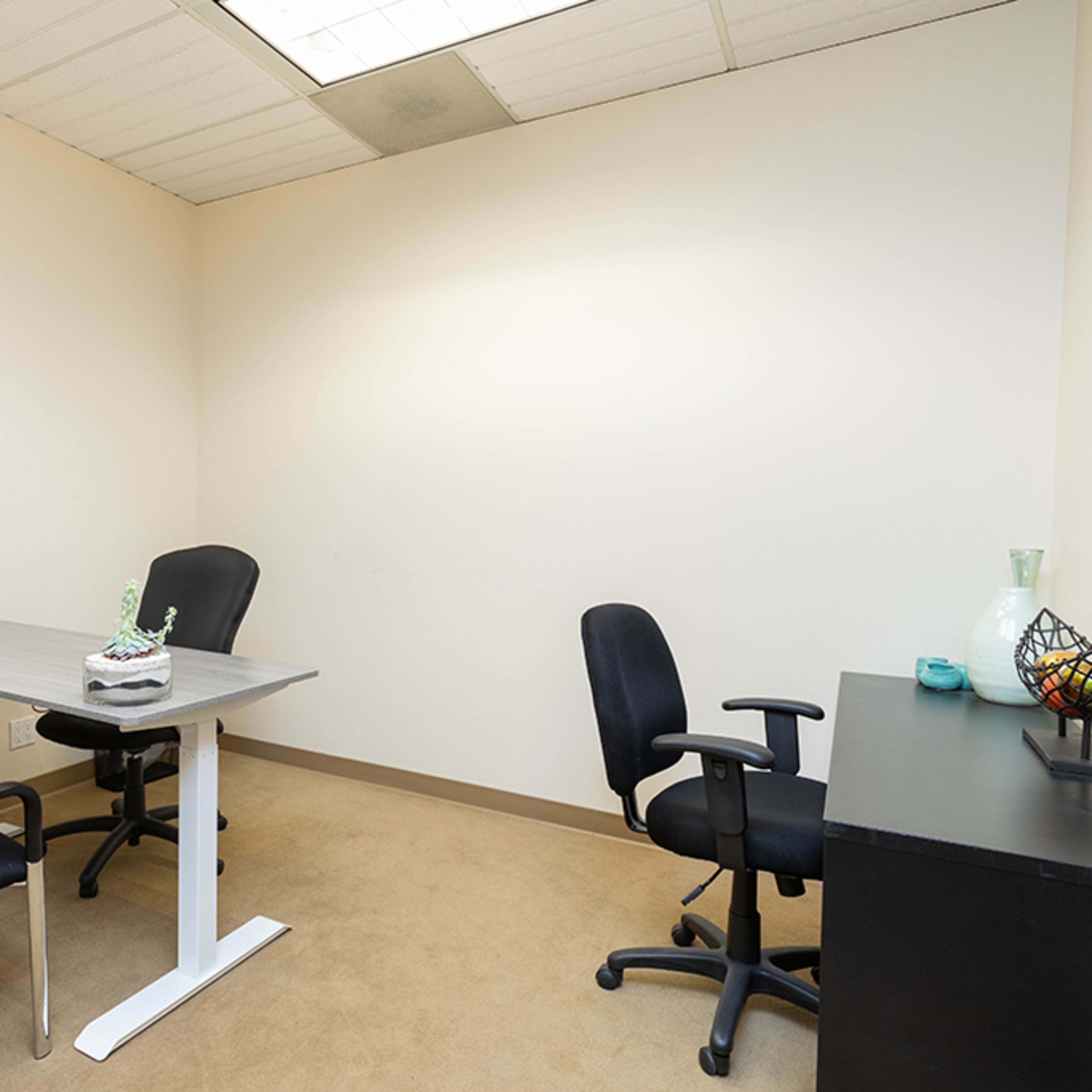 An empty office features a desk with a phone and a small decorative item, four chairs, and a black cabinet along the wall.