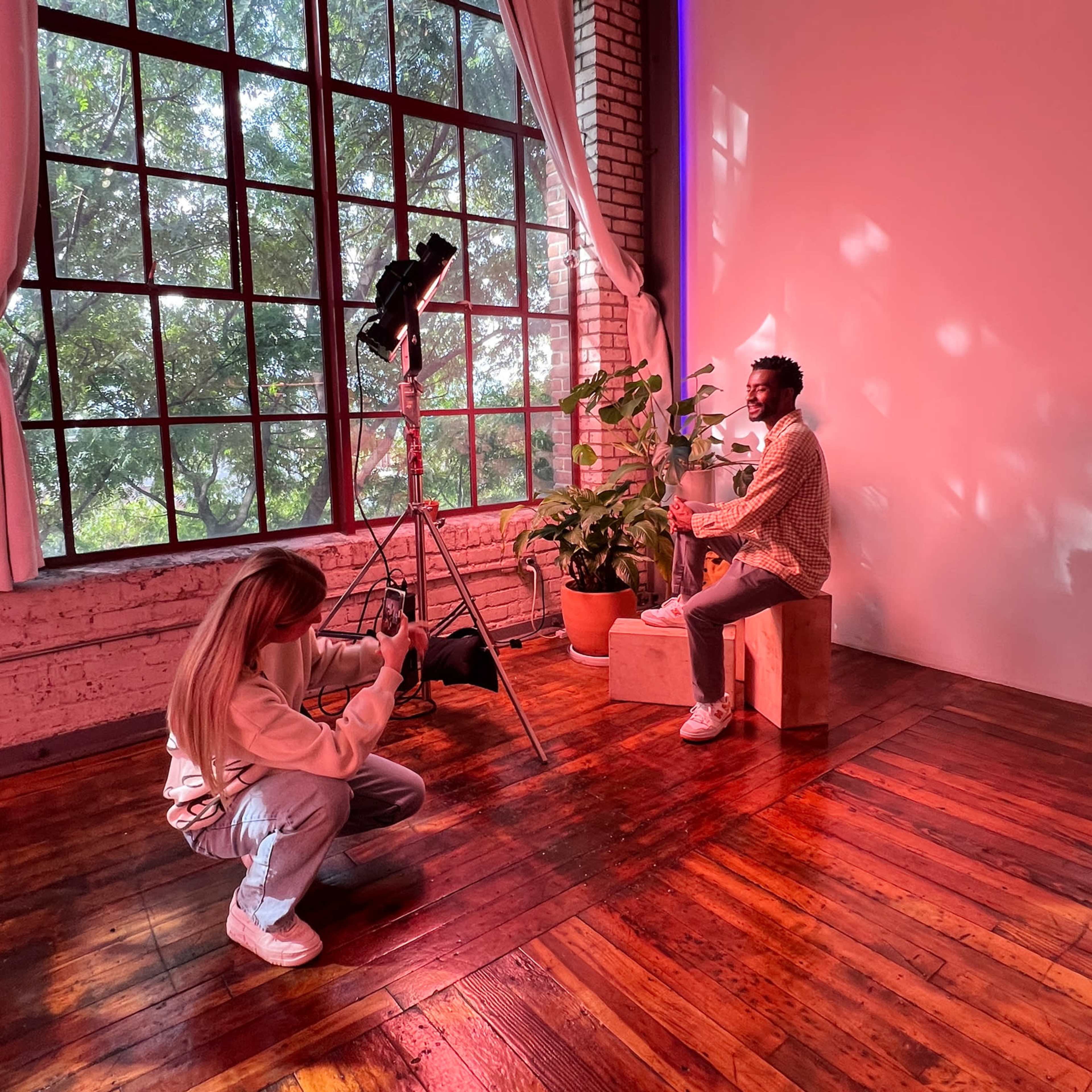 A woman operates a camera while a man poses on a wooden box in front of a large window adorned with greenery.