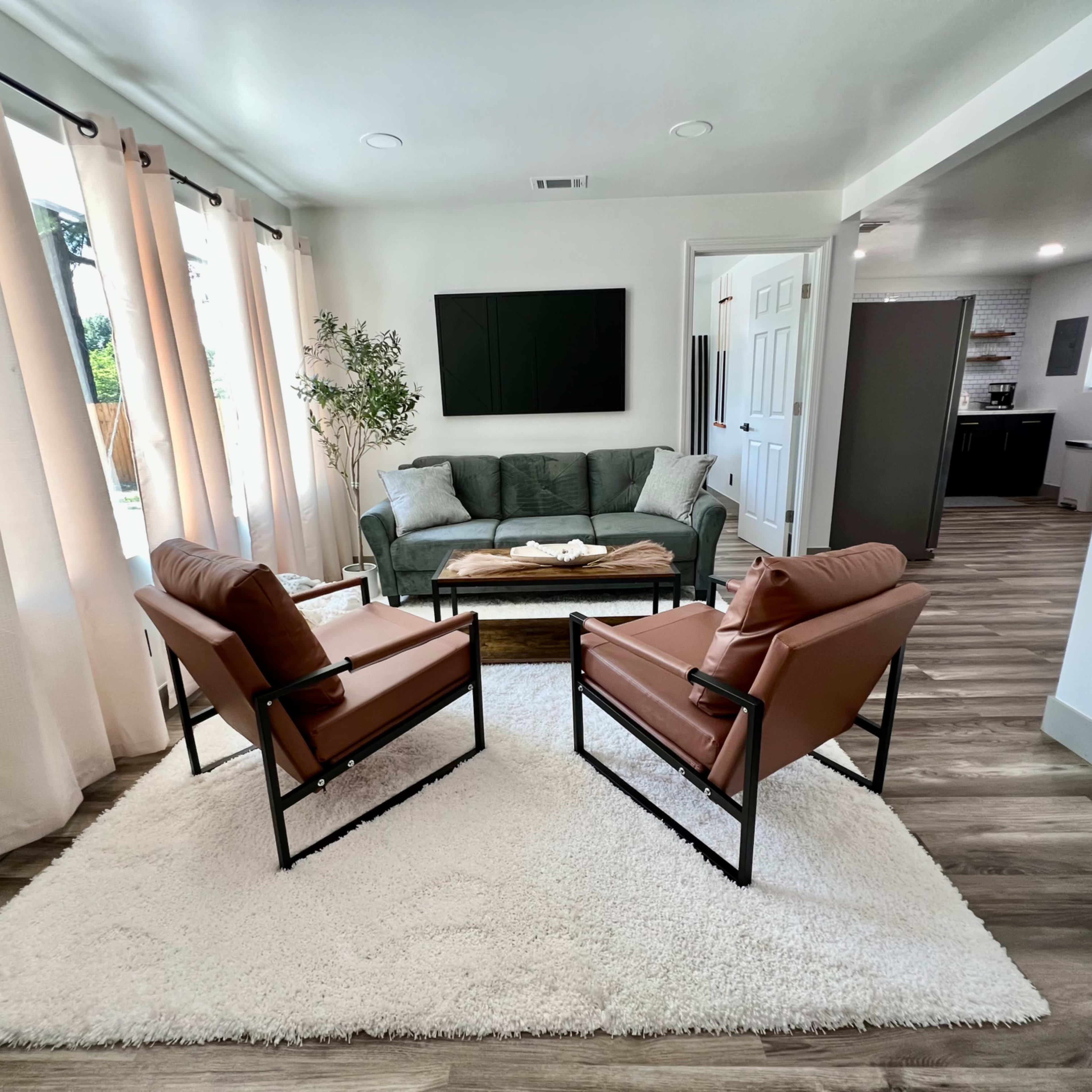 A modern living room with a gray sofa, two brown chairs, a coffee table, and large windows dressed with light curtains.