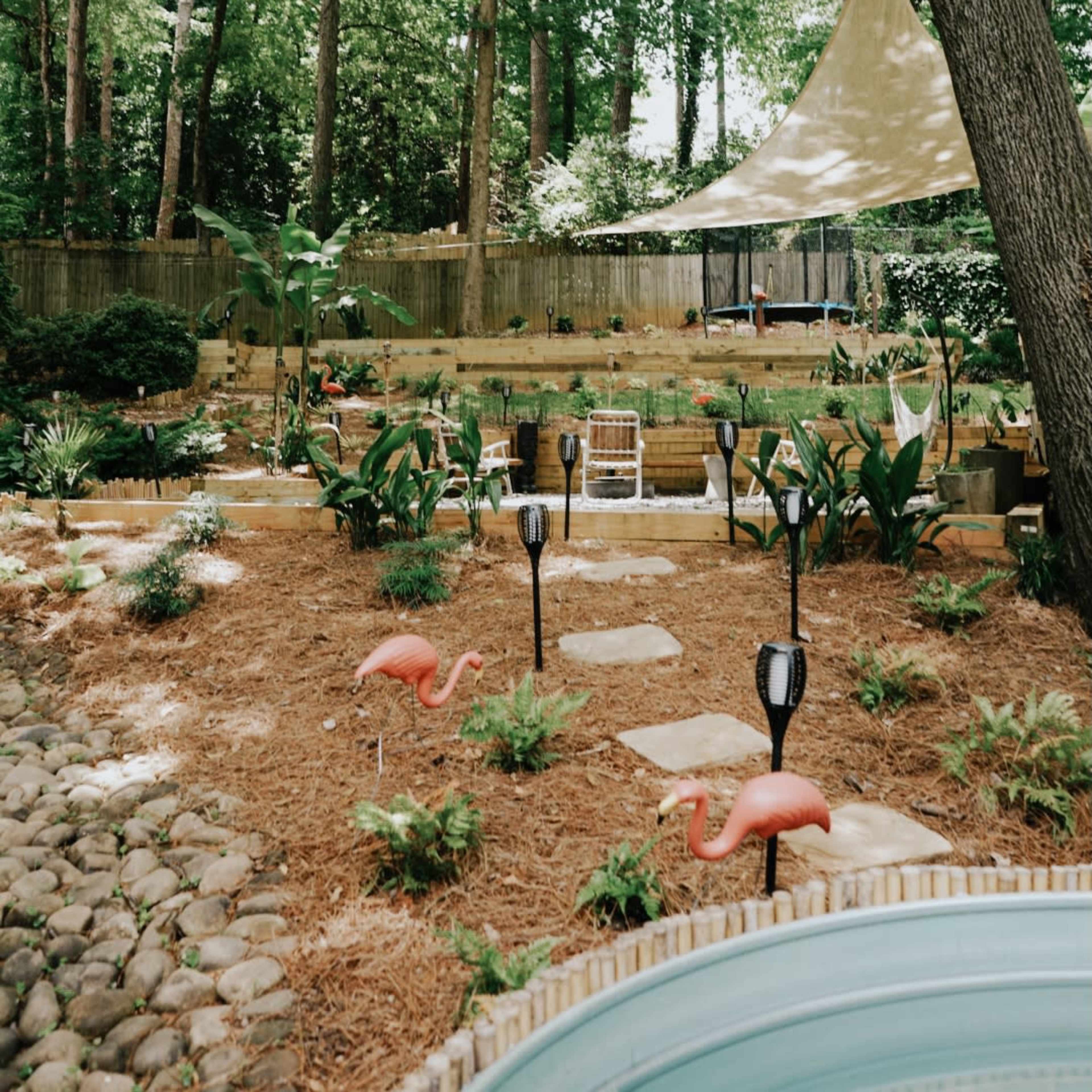 A landscaped garden with a stone pathway, surrounded by greenery, featuring pink flamingo decorations and a shaded area overhead.