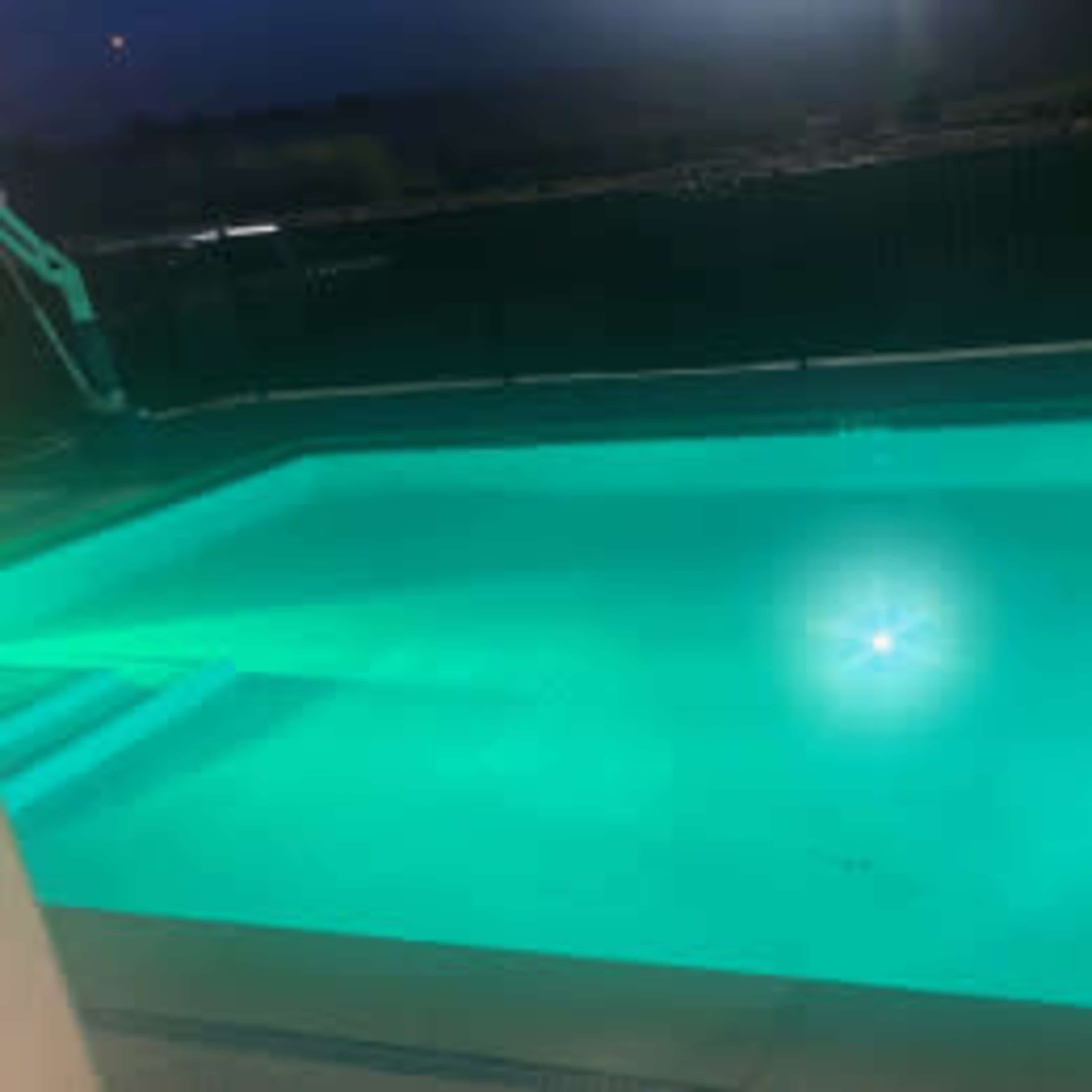 A brightly lit swimming pool at night, with a glowing blue interior and a ladder on the side.