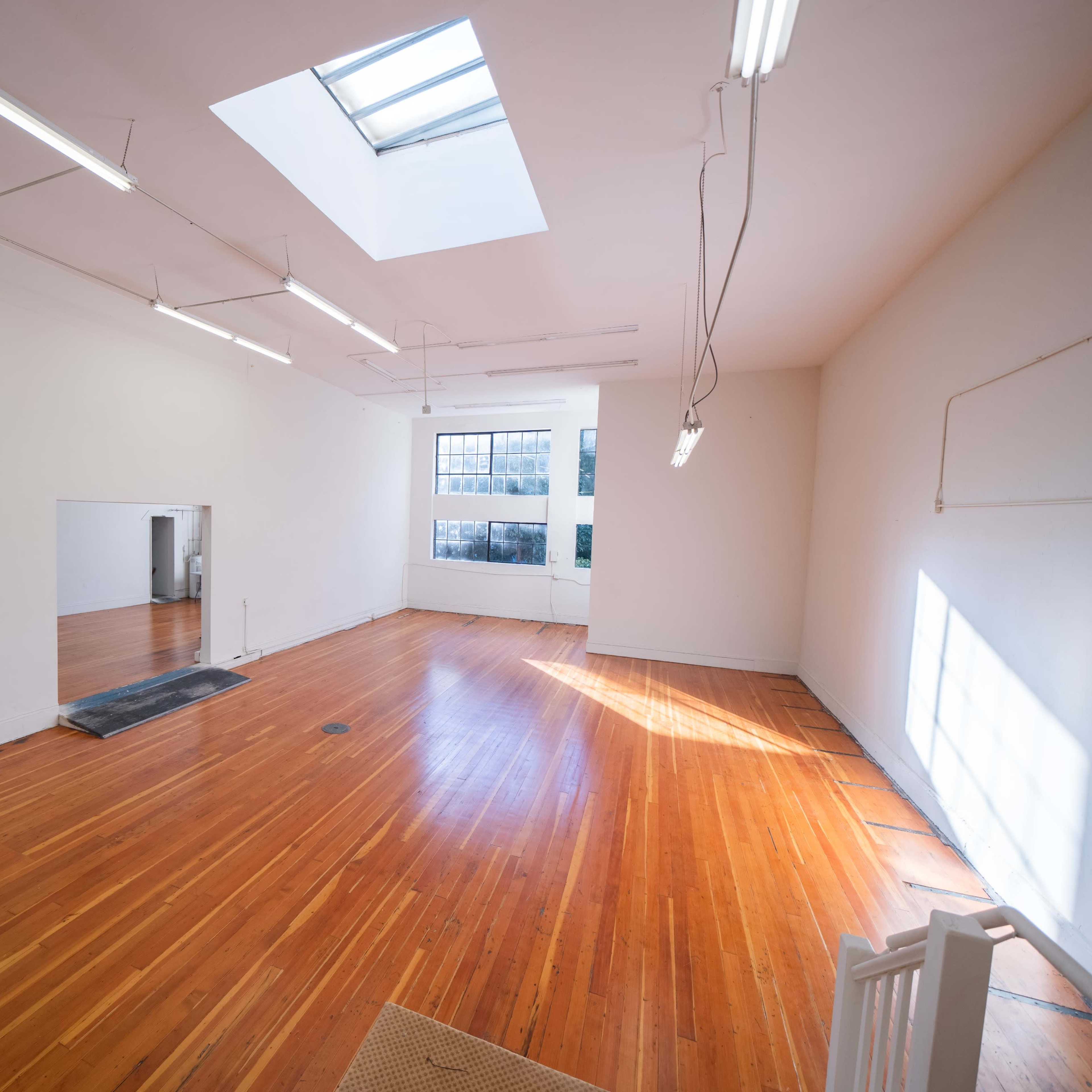 A spacious, empty room with hardwood floors, a skylight, and a large window, illuminated by fluorescent lights.