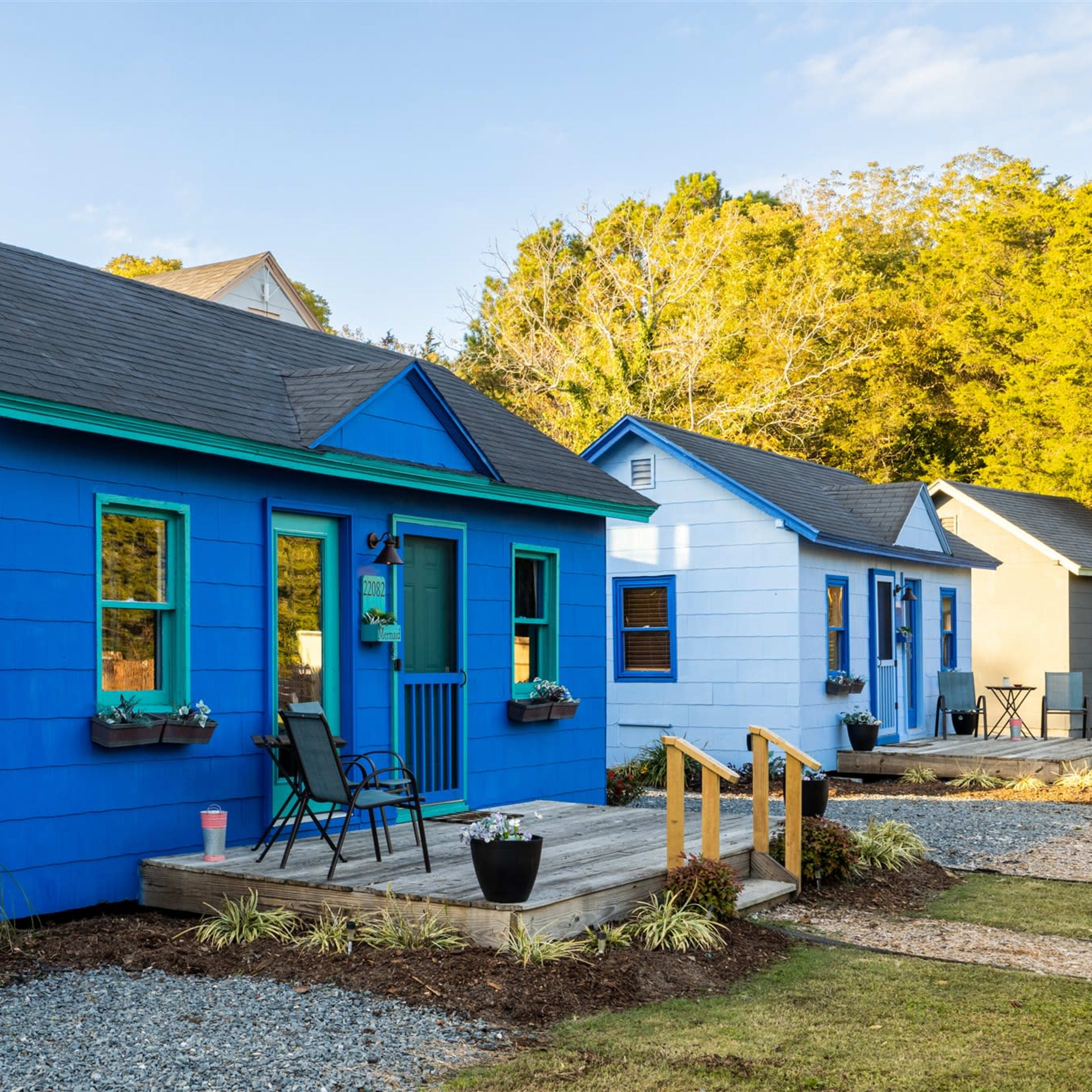 Two brightly colored homes, one blue and one light blue, are situated side by side with a wooden porch and steps in a landscaped setting.