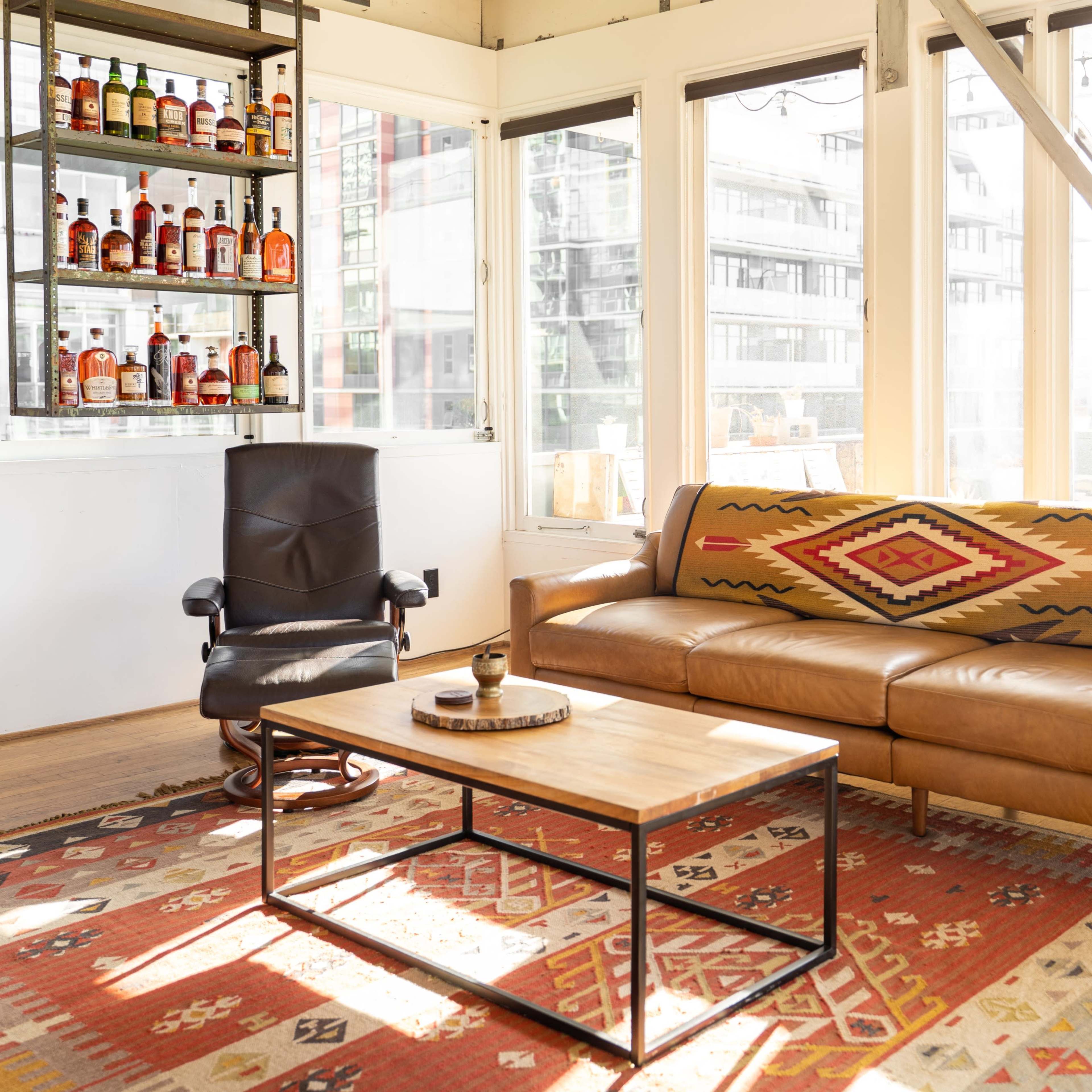 A modern living room features a brown leather sofa with a patterned throw, a wooden coffee table, and a black rocking chair, surrounded by large windows and a shelf displaying various bottles.