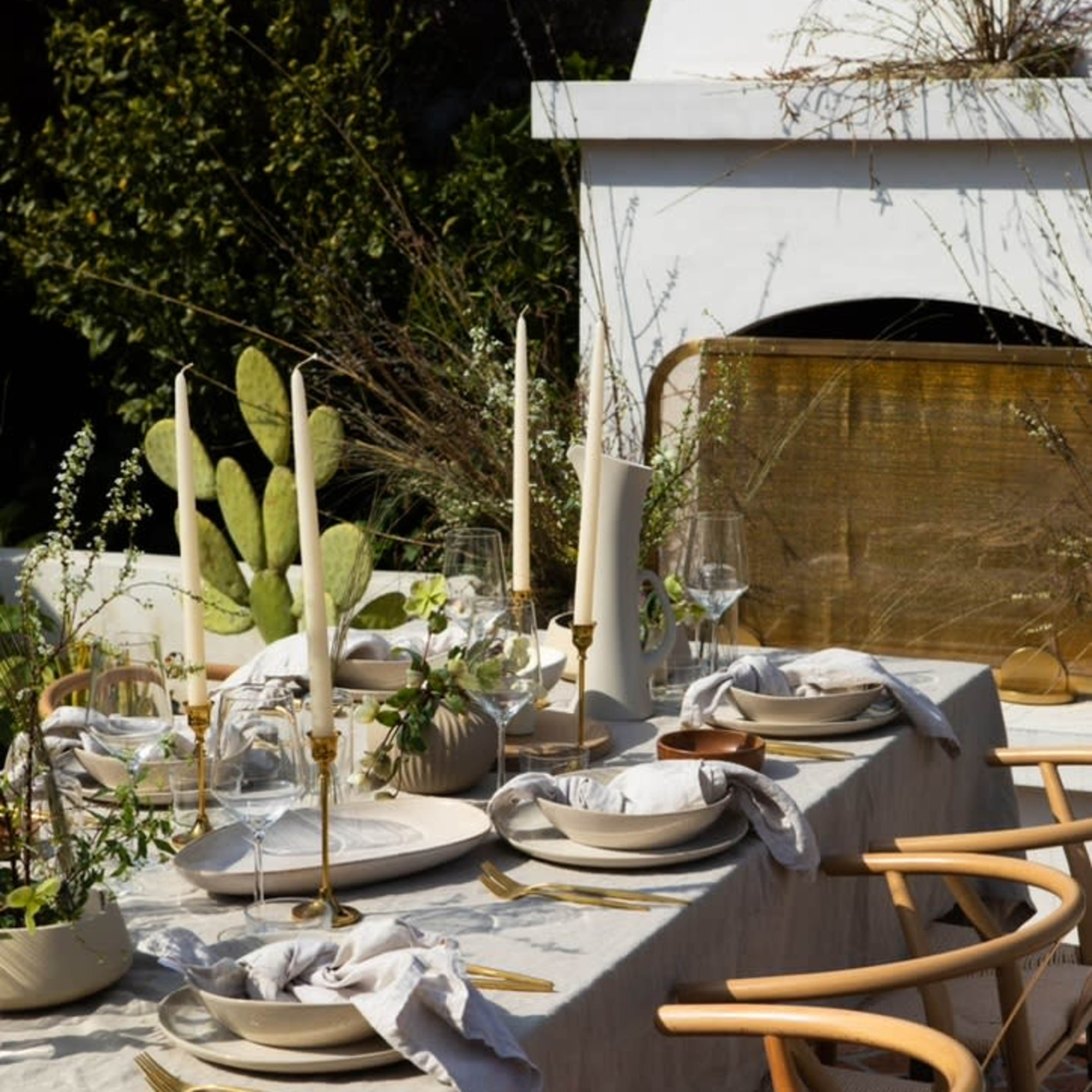 A long, elegantly set dining table features white plates, glassware, and candles, surrounded by wooden chairs in an outdoor setting with greenery.