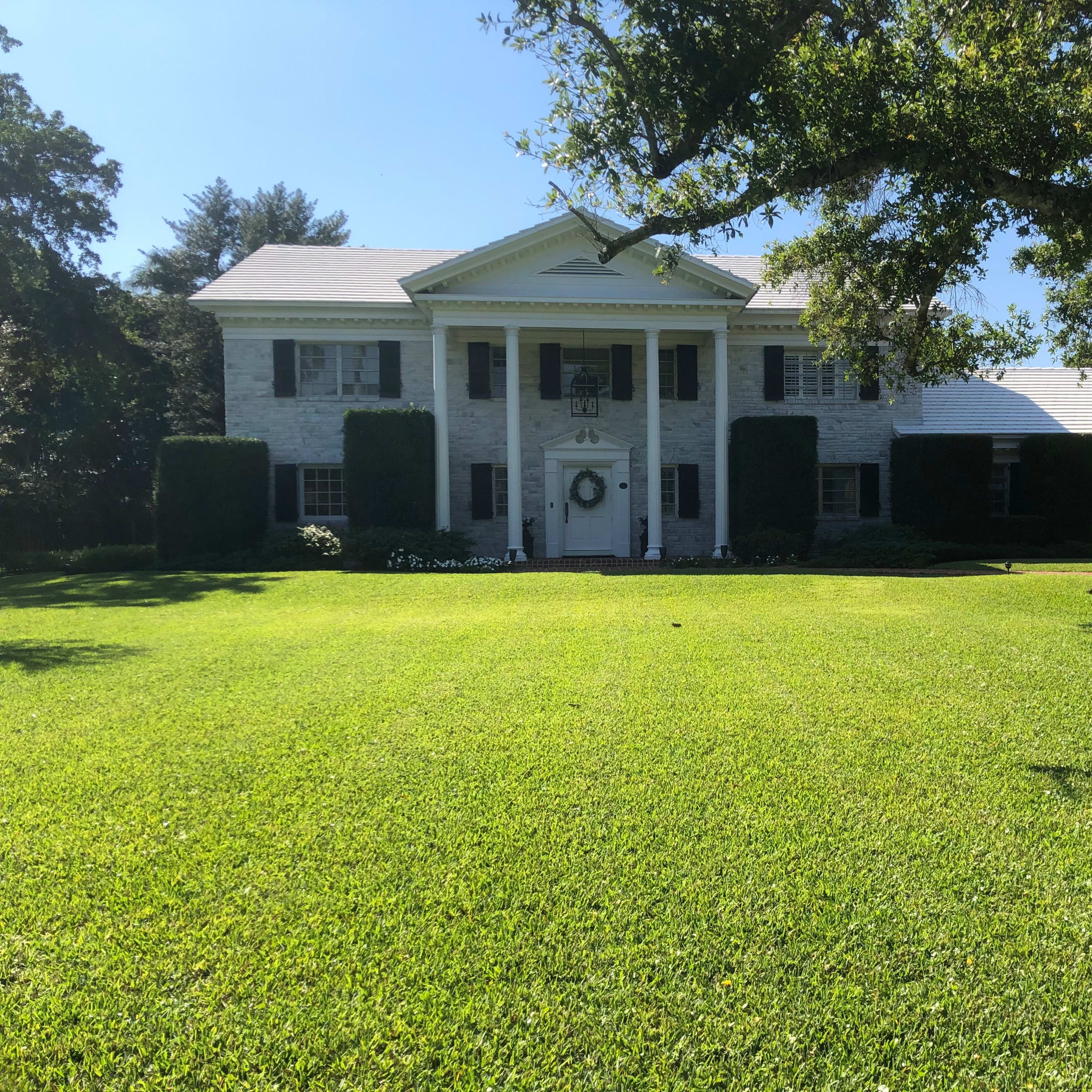 A large, two-story house with white stone exterior and black shutters sits on a well-manicured lawn surrounded by neatly trimmed bushes and trees.