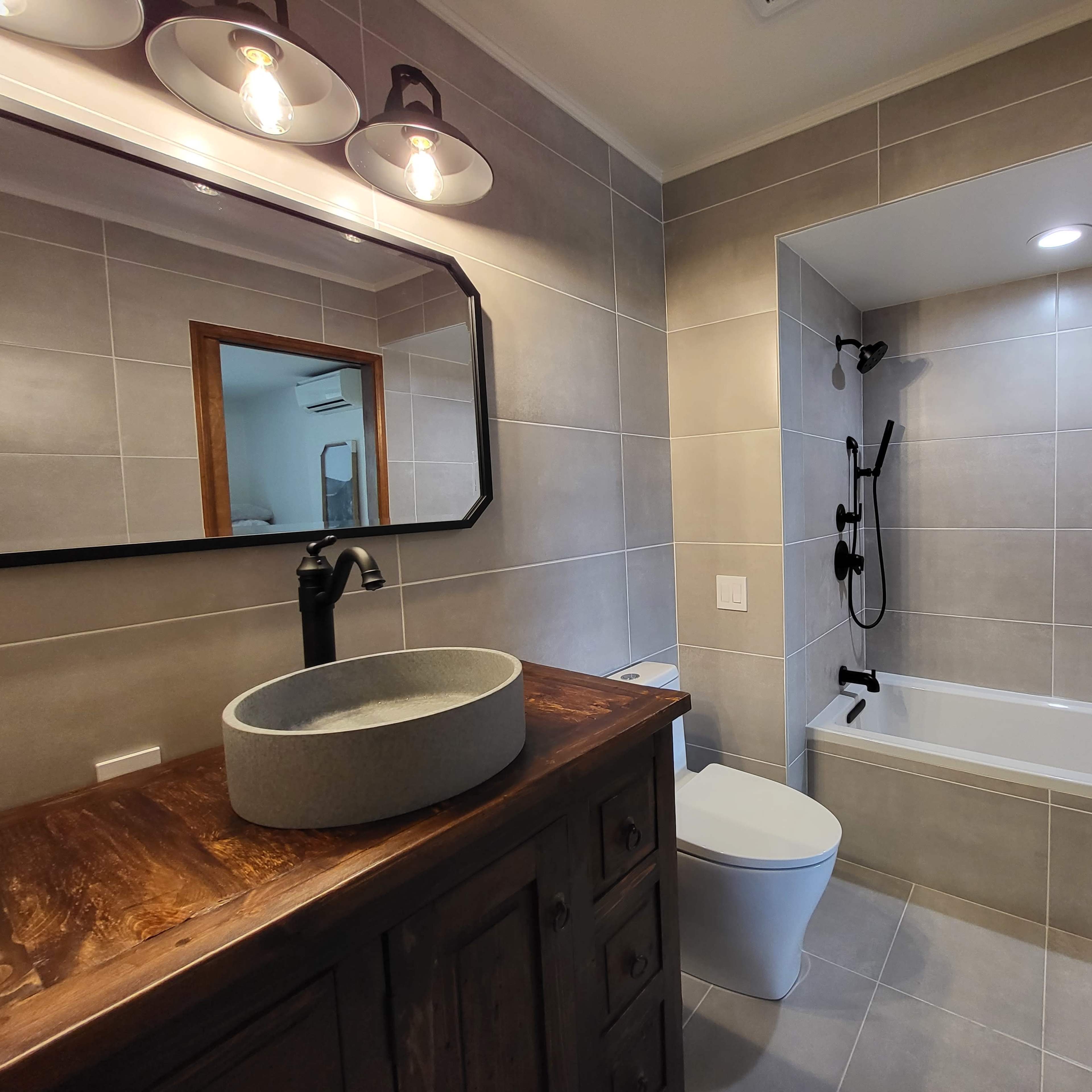 A modern bathroom with a wooden vanity, a round sink, a white toilet, a bathtub, and a tiled wall with a mirror and light fixtures.