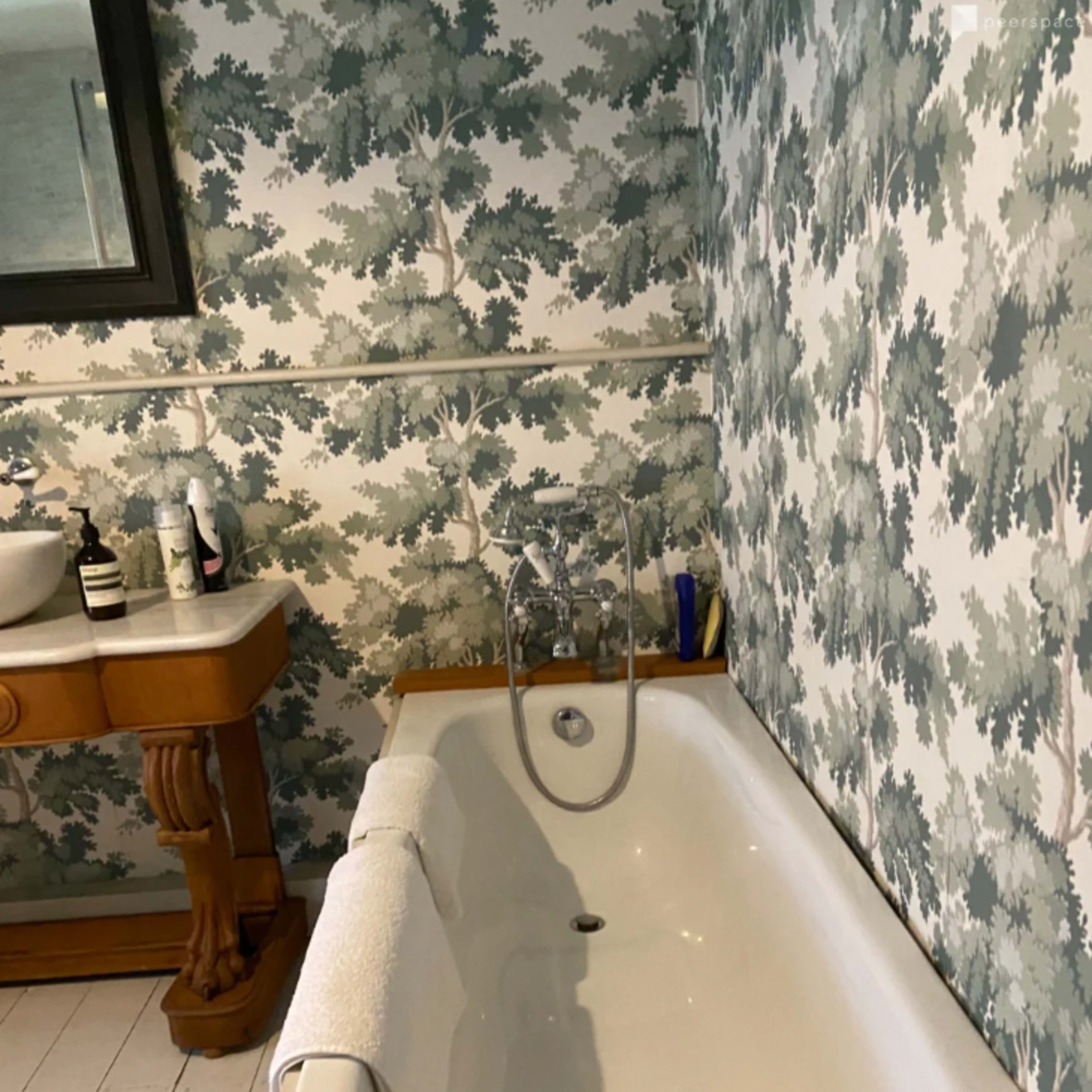 A white bathtub with a towel draped over it is positioned next to a wooden vanity set against a wall covered in green and white tree-patterned wallpaper.