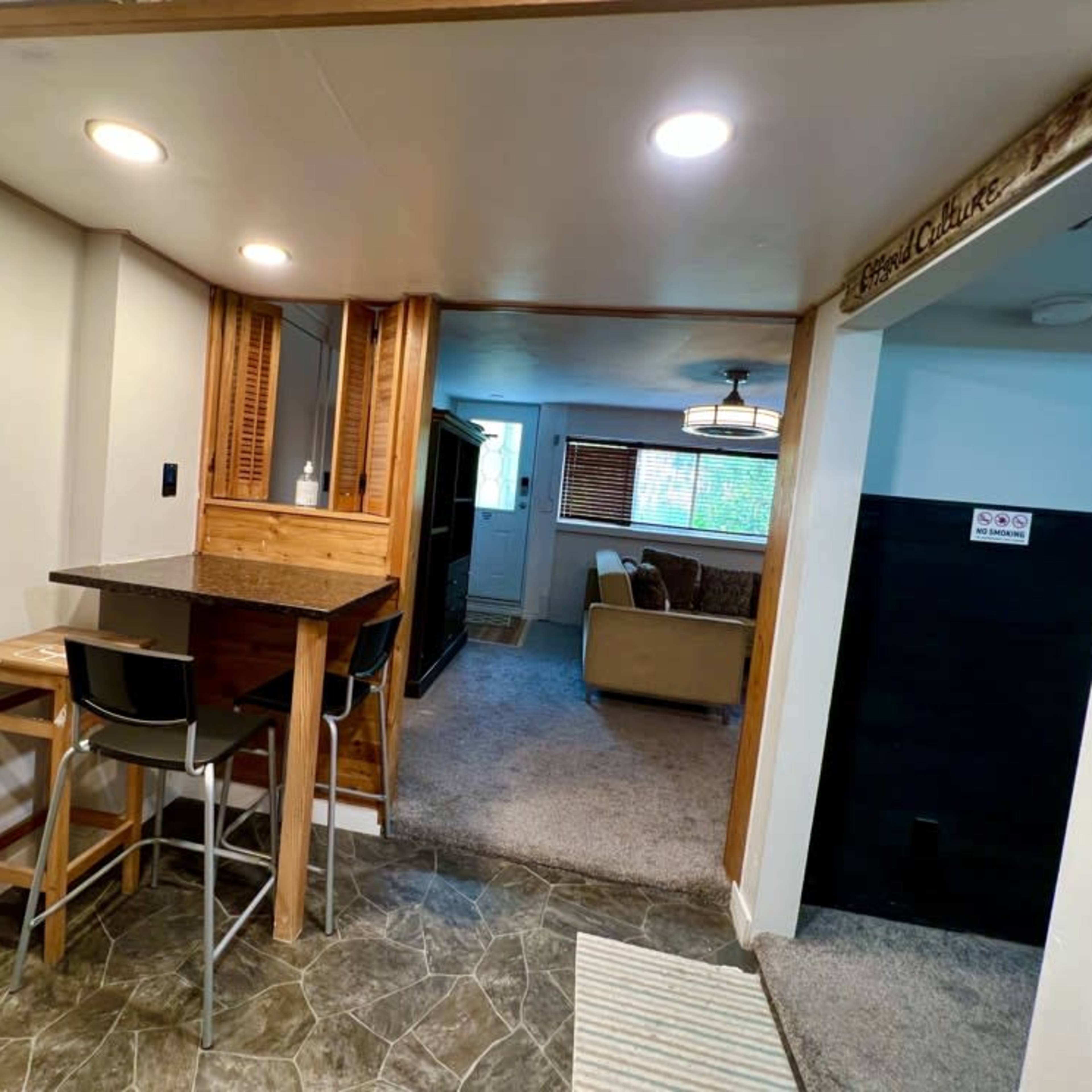 A small kitchen area with a bar table and two stools, leading into a carpeted living room with a sofa and window.