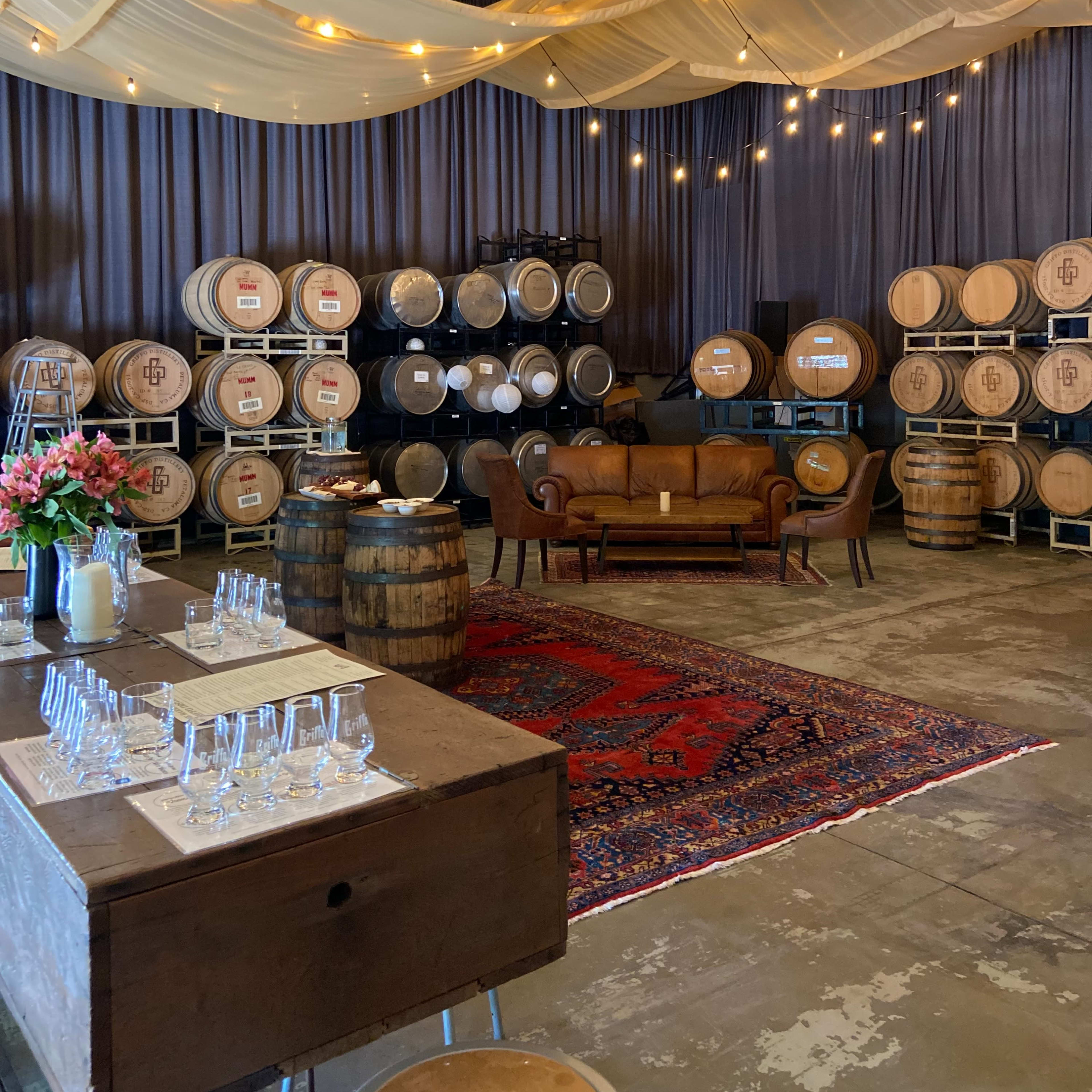 A spacious room features wooden barrels stacked against a wall, a rustic couch, and a decorated table with glassware, all under string lights and draped fabric.
