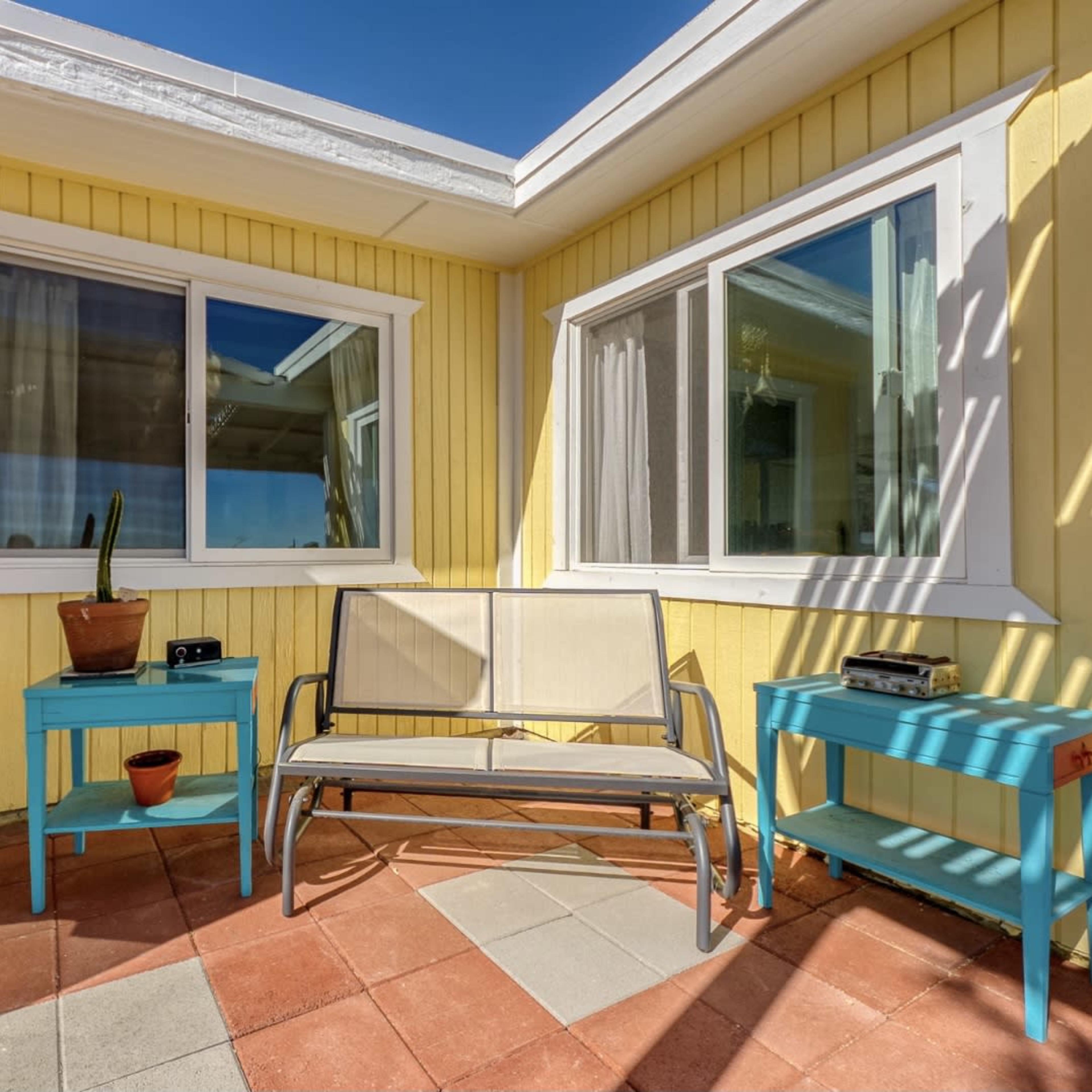 A small patio area features a beige bench flanked by two turquoise side tables, with potted plants and large windows set into a yellow wall.
