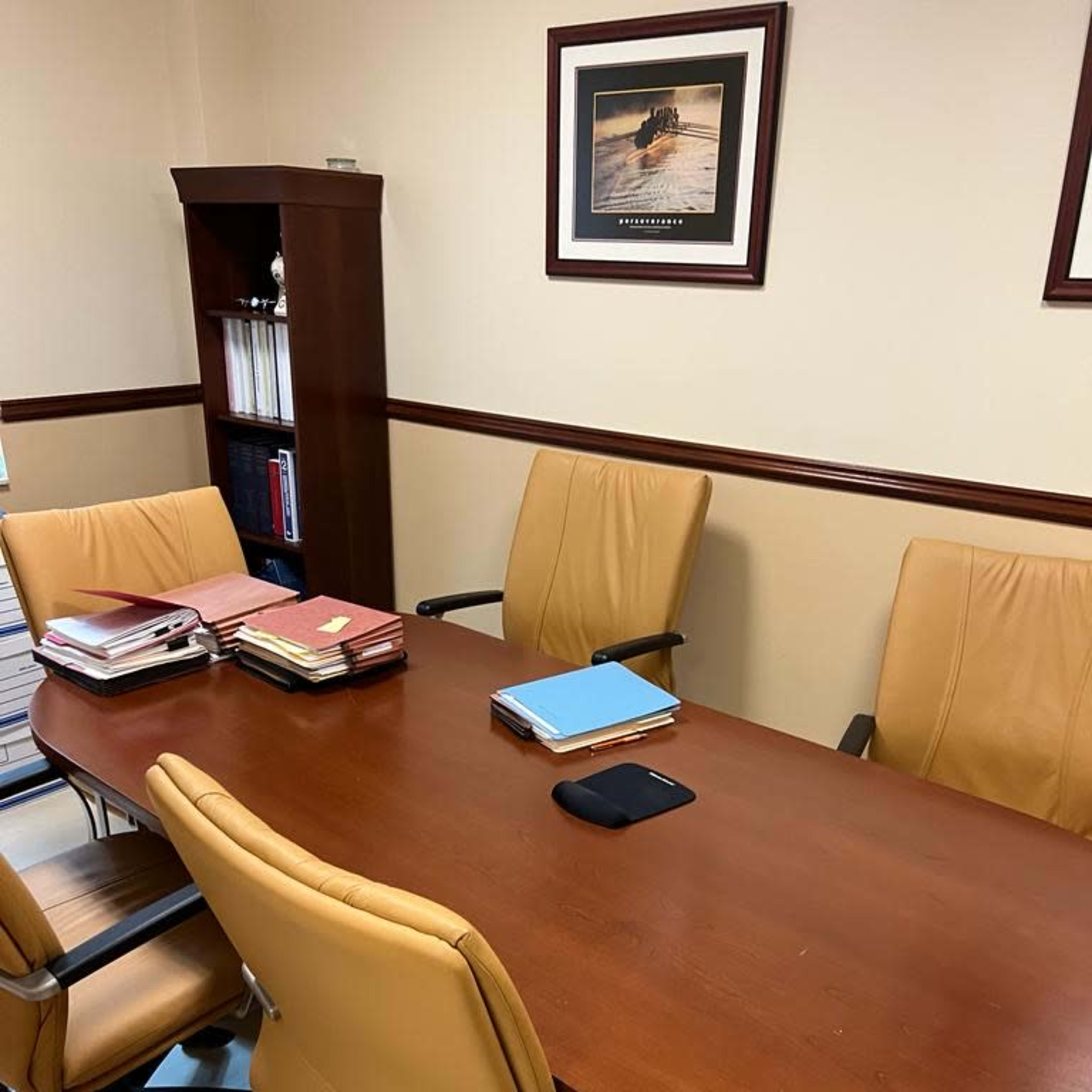 A wooden conference table surrounded by four tan office chairs is shown in a well-lit office with a framed picture on the wall and a bookshelf in the corner.