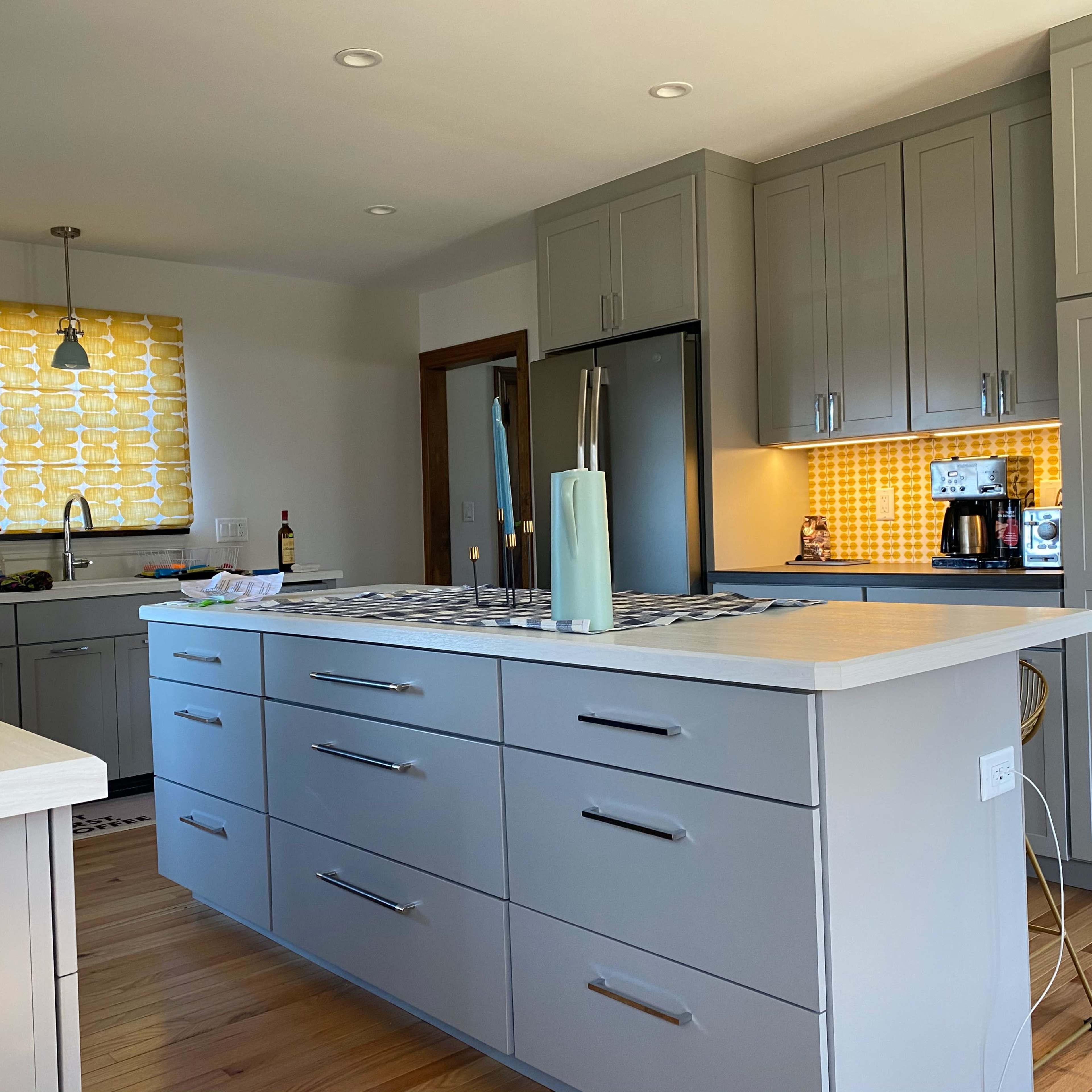 A modern kitchen features gray cabinets, a central island with a light countertop, and a window with a yellow patterned shade.