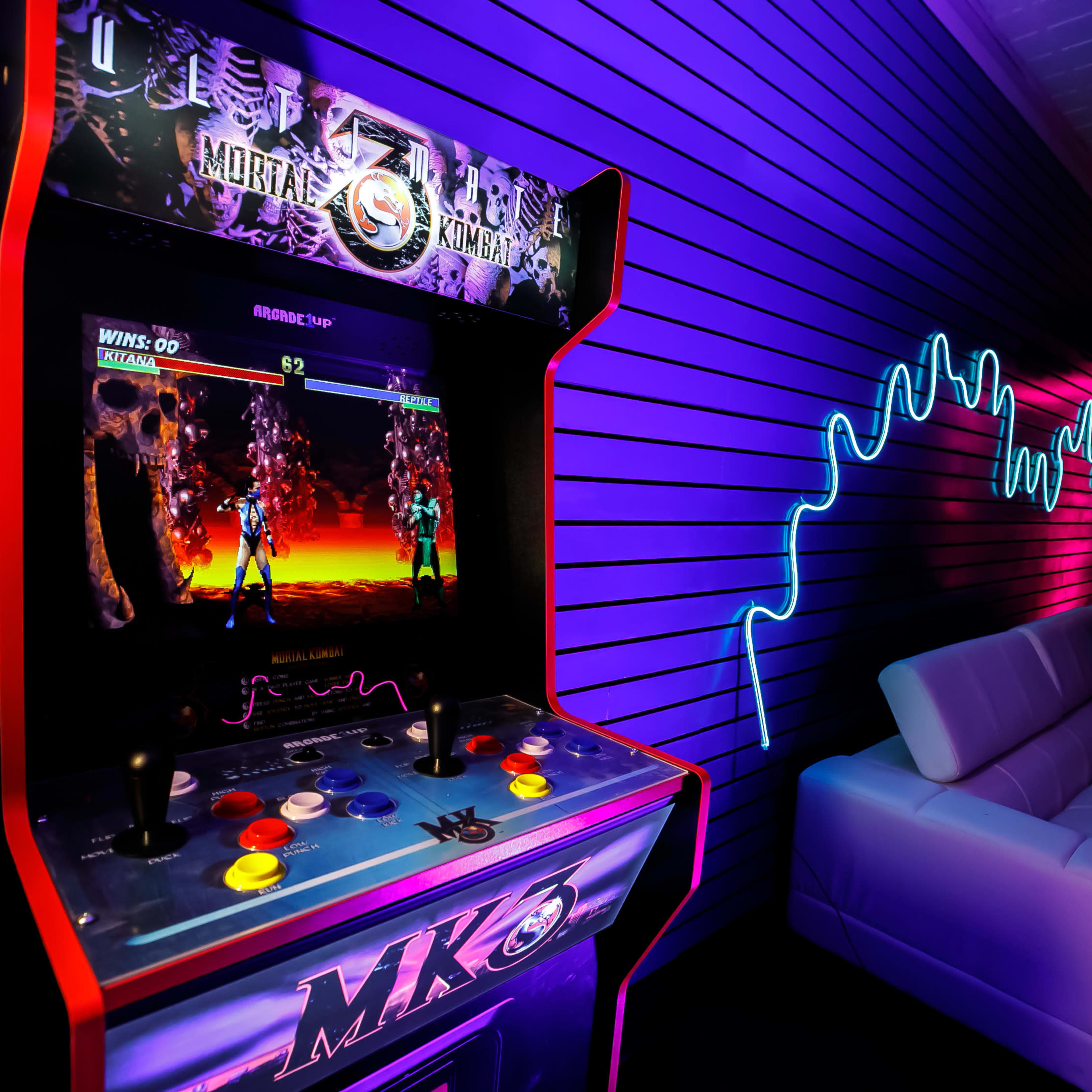 A retro arcade machine displaying a game of Mortal Kombat against a backdrop of colorful neon lights and a modern white couch.