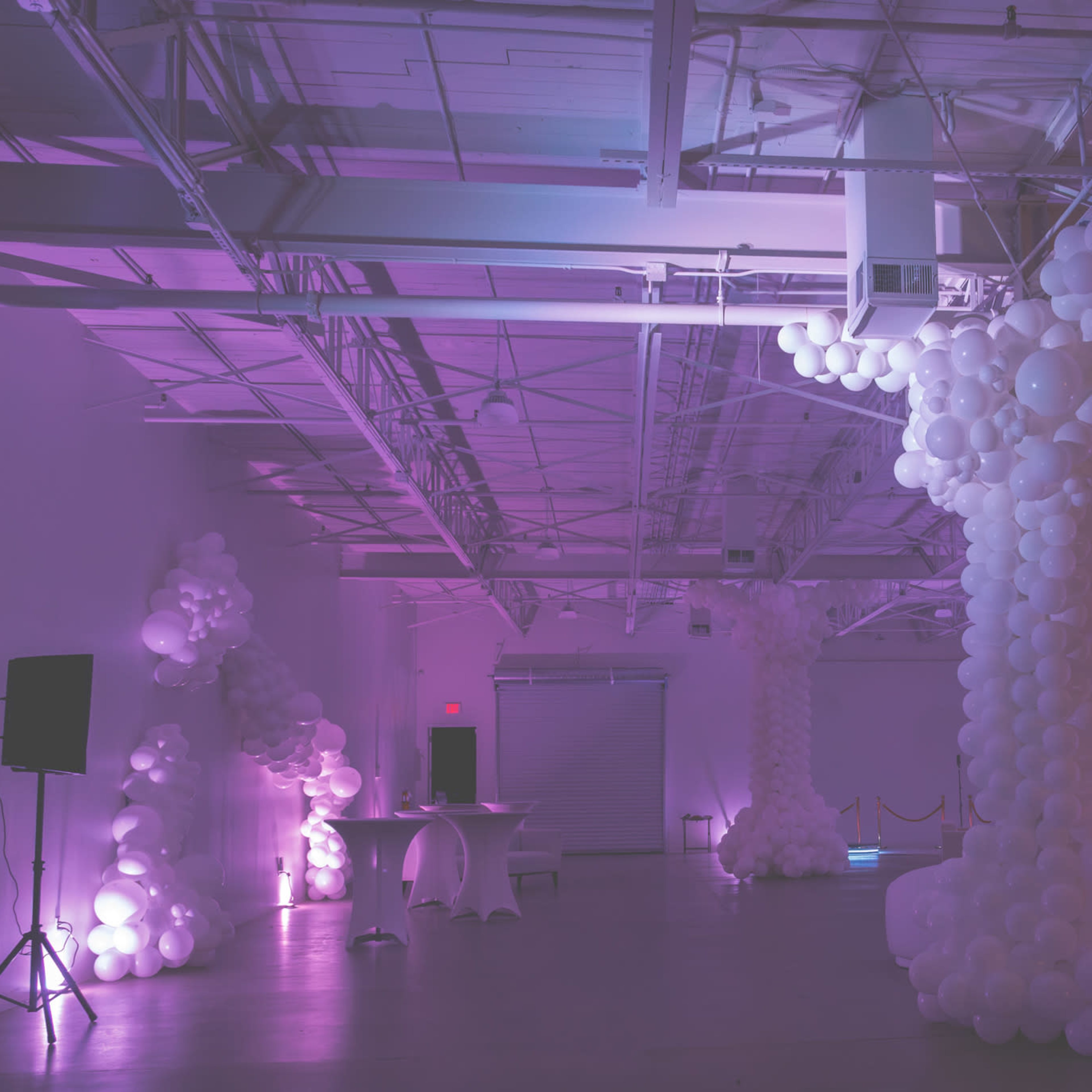 A spacious event venue decorated with large white balloon installations and dim purple lighting.