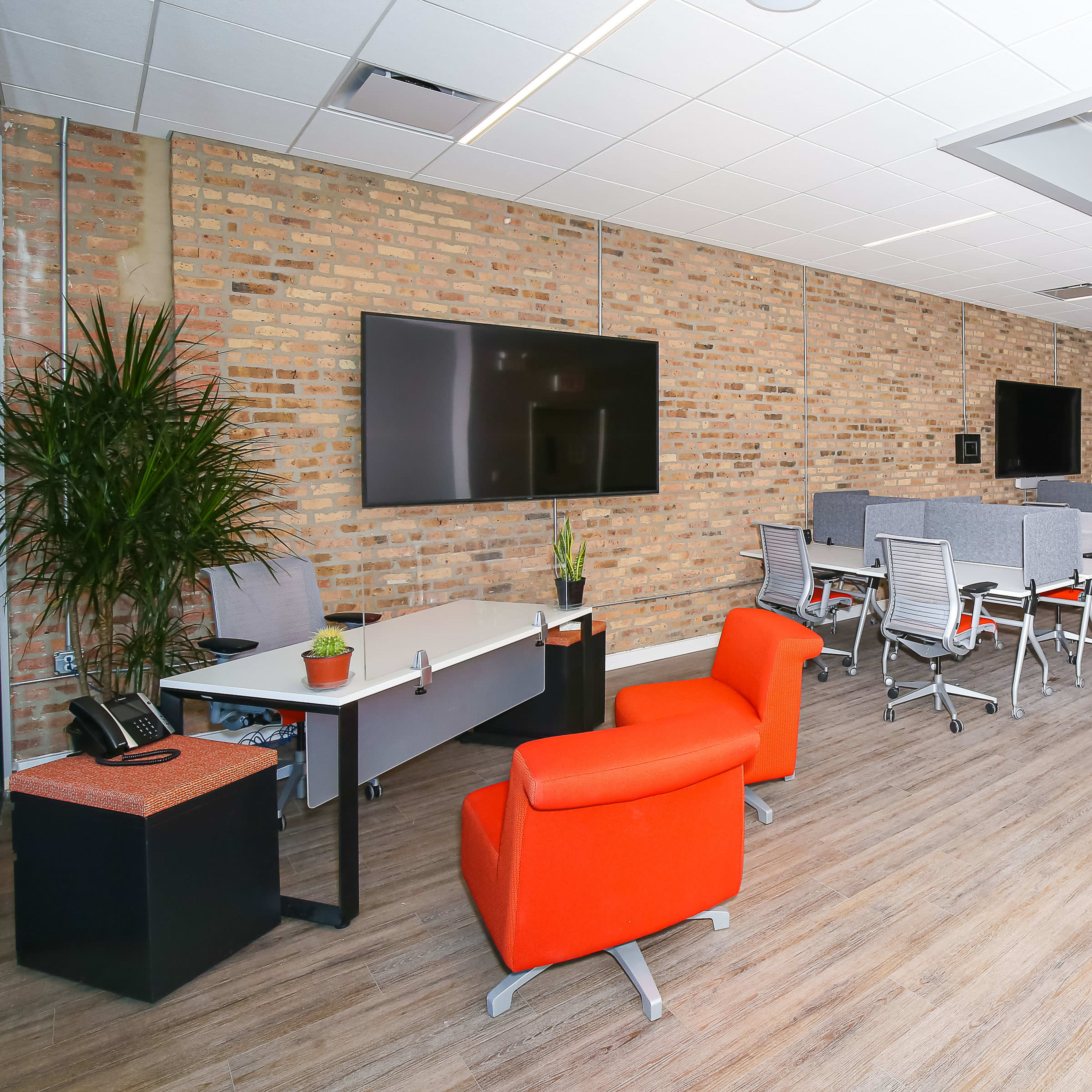 A spacious office area with a mix of workstations, a seating area, and two large television screens mounted on a brick wall.