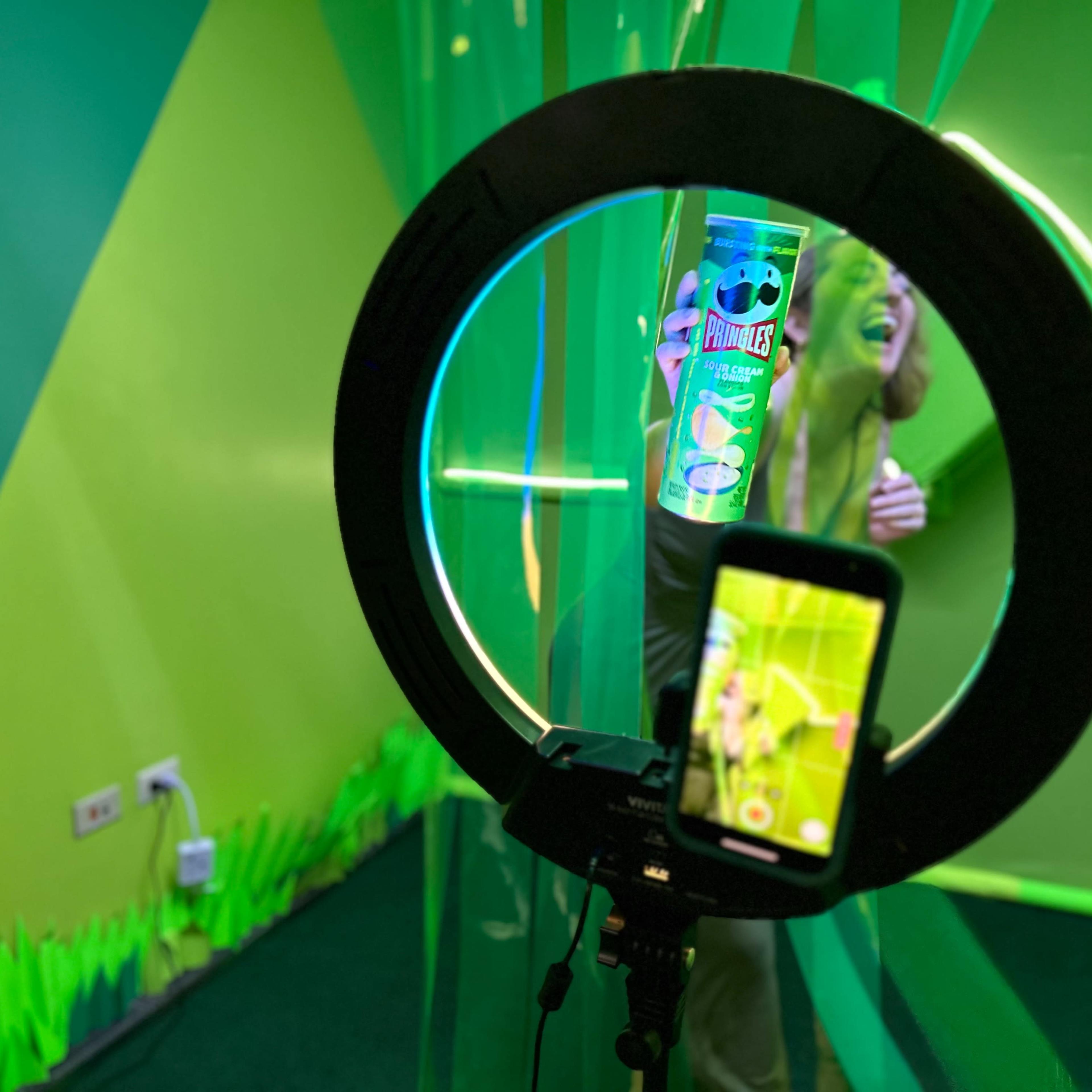 A circular ring light frames a can of Pringles while a person interacts with green streamers in a brightly colored room.