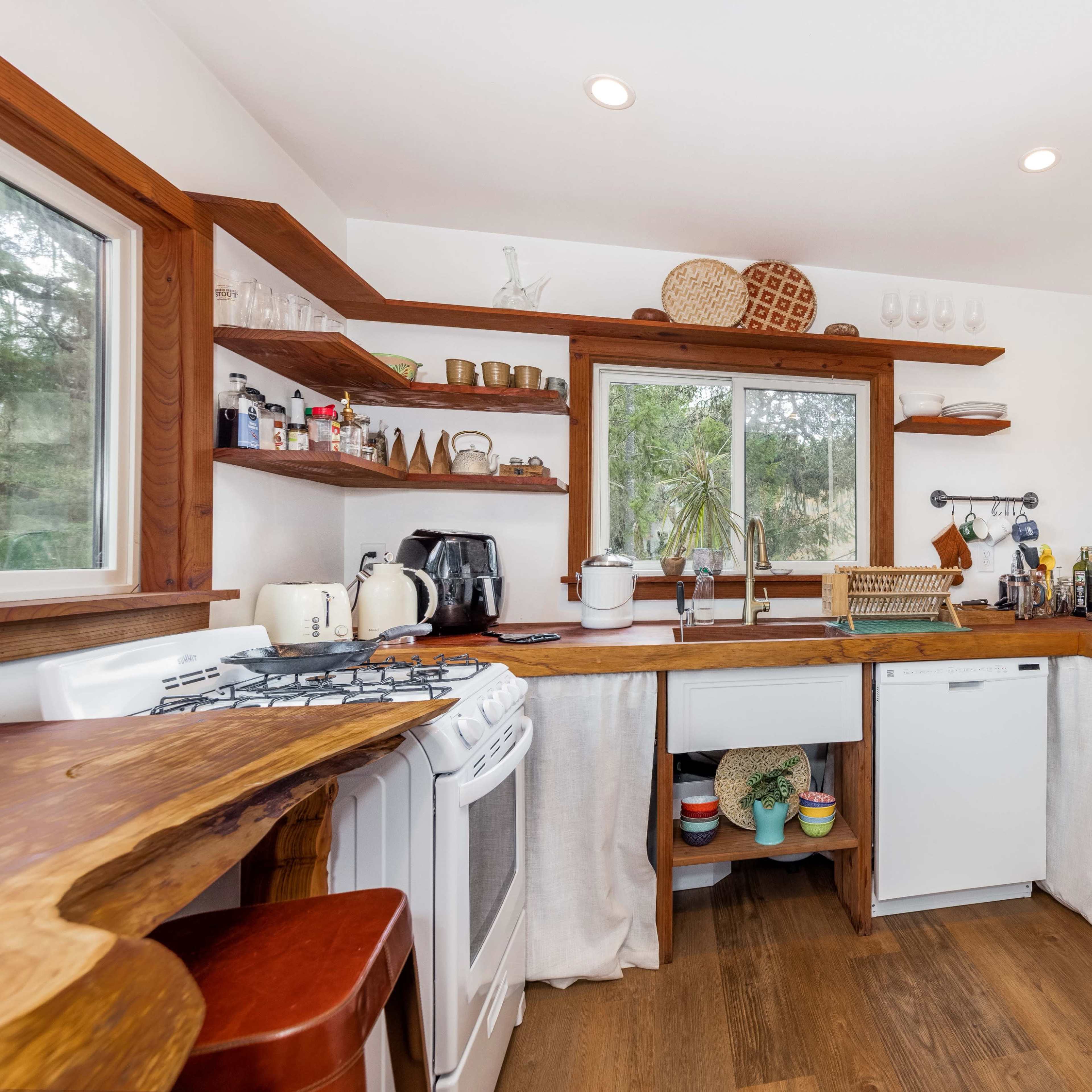 A compact kitchen features wooden shelves, a white refrigerator, and a small dining area with a view of the outdoors.