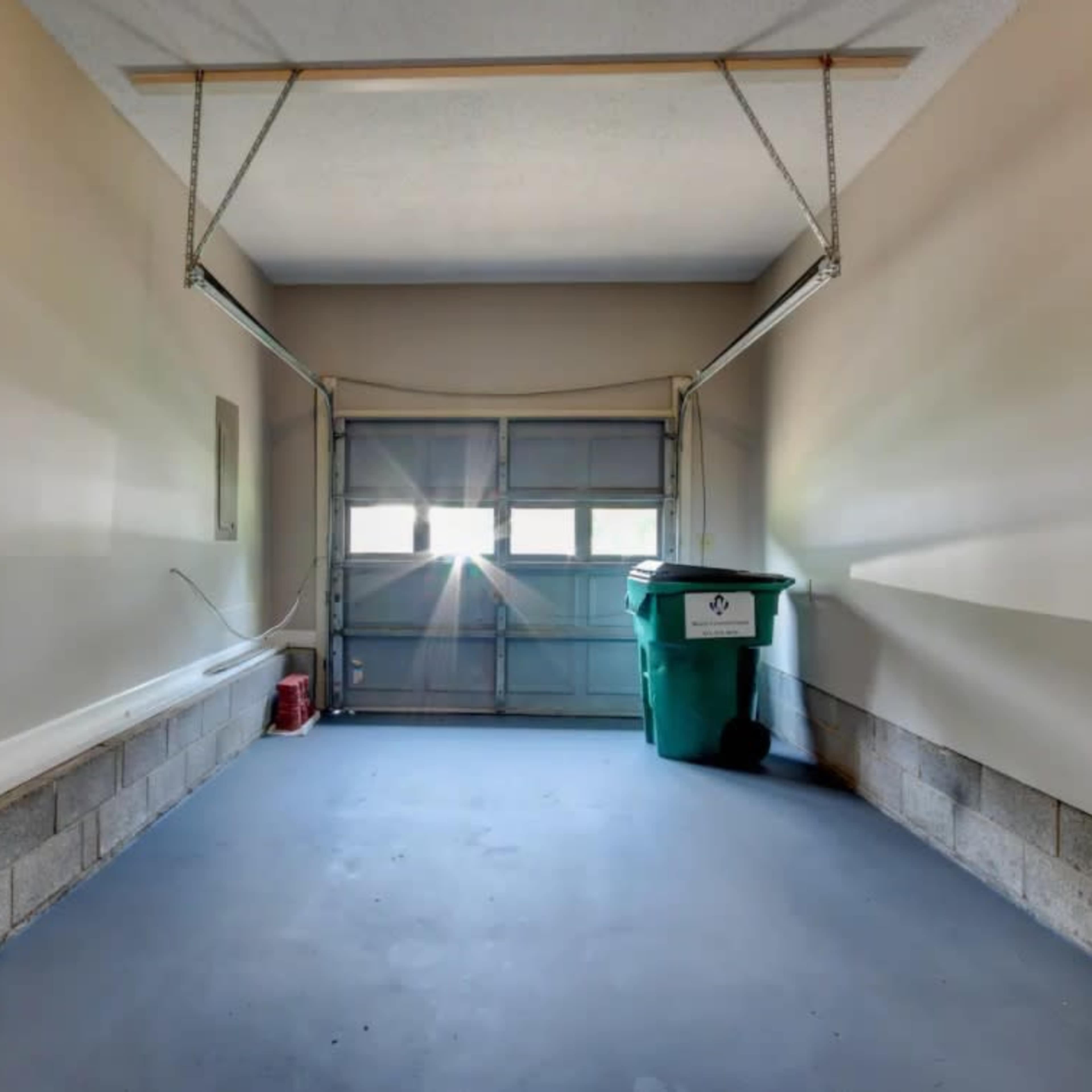 An empty garage with a green trash bin near the closed garage door, where sunlight is streaming in through the windows.