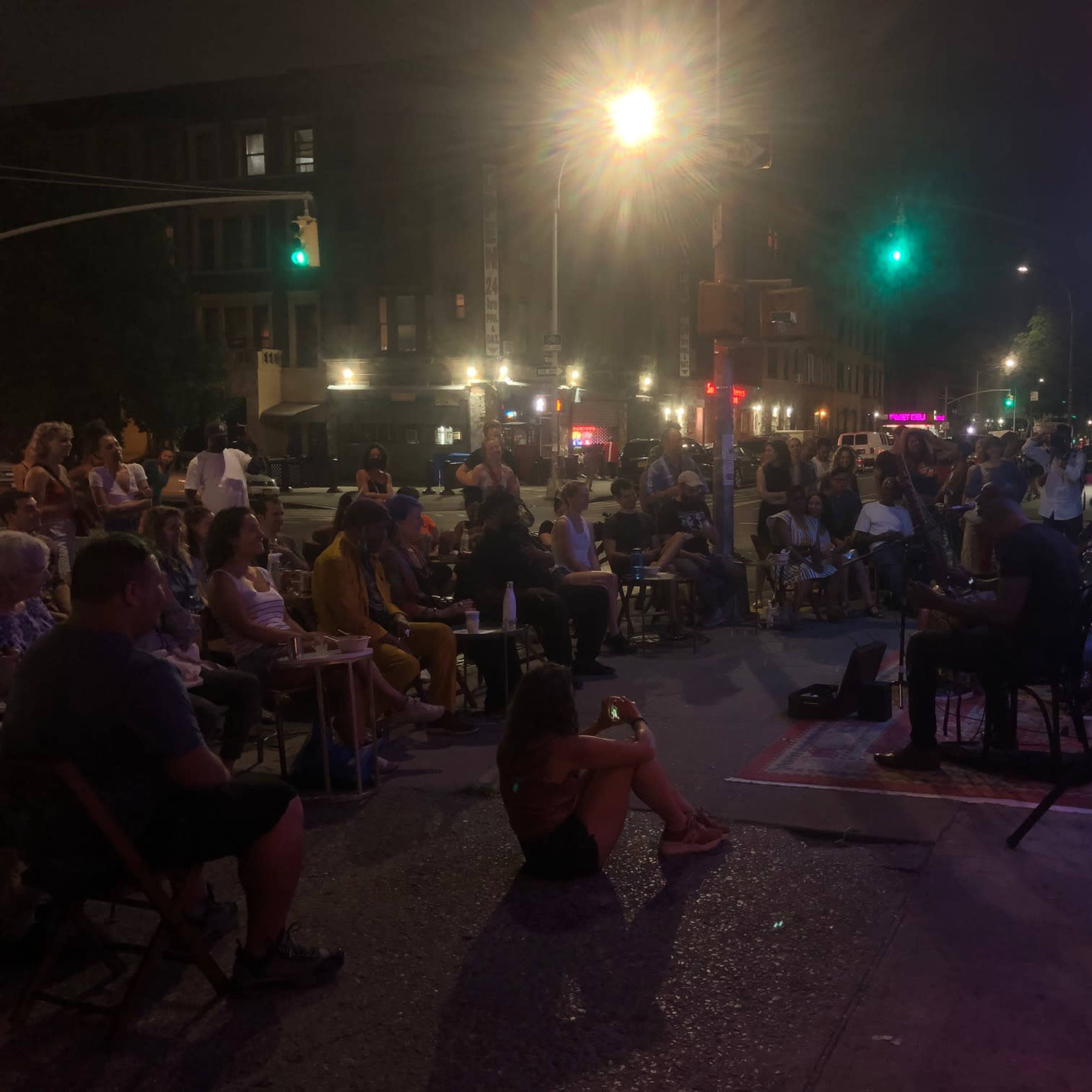 A crowd sits on the street, enjoying a live music performance at night.