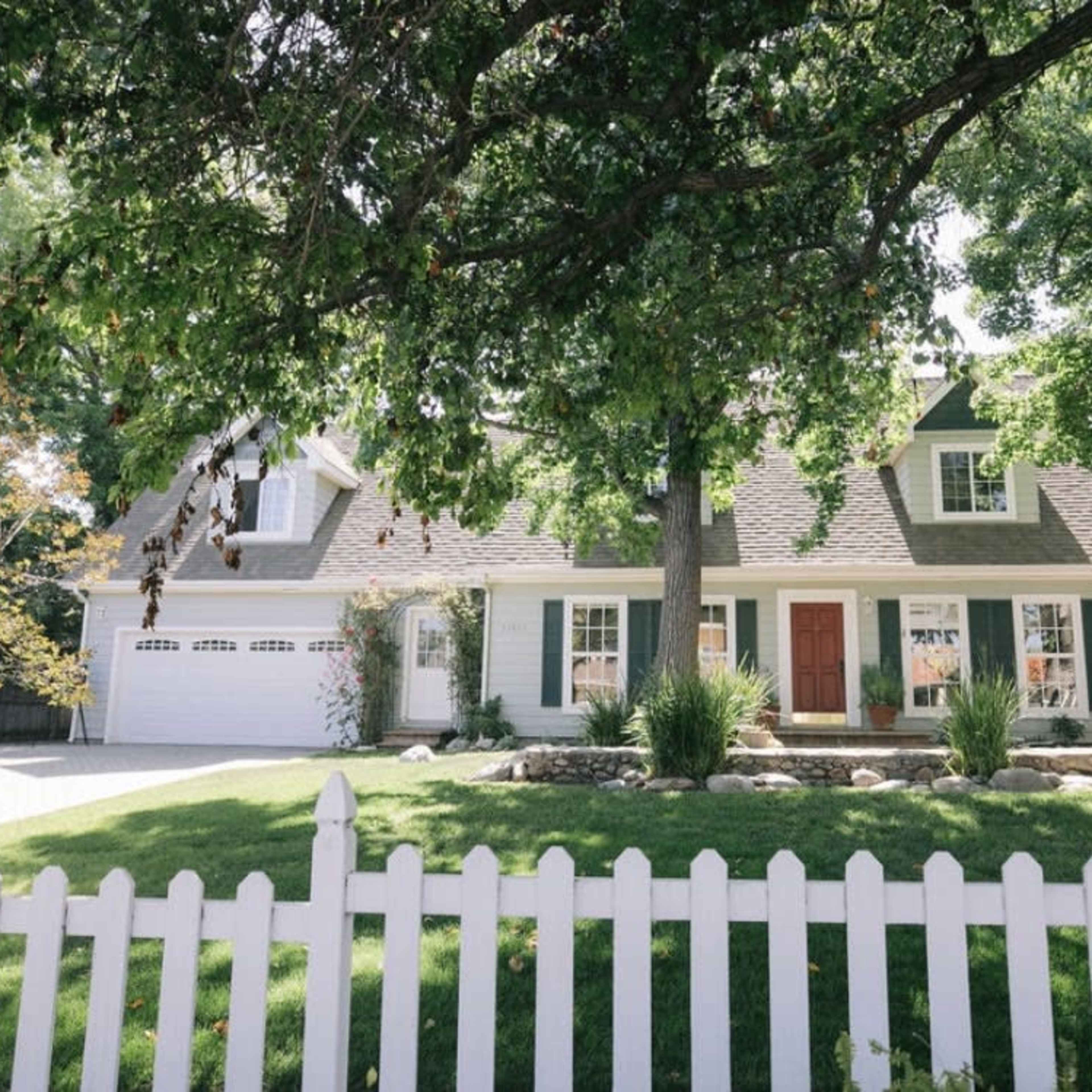 A white picket fence surrounds a green lawn in front of a two-story house with a gabled roof and a large front porch.