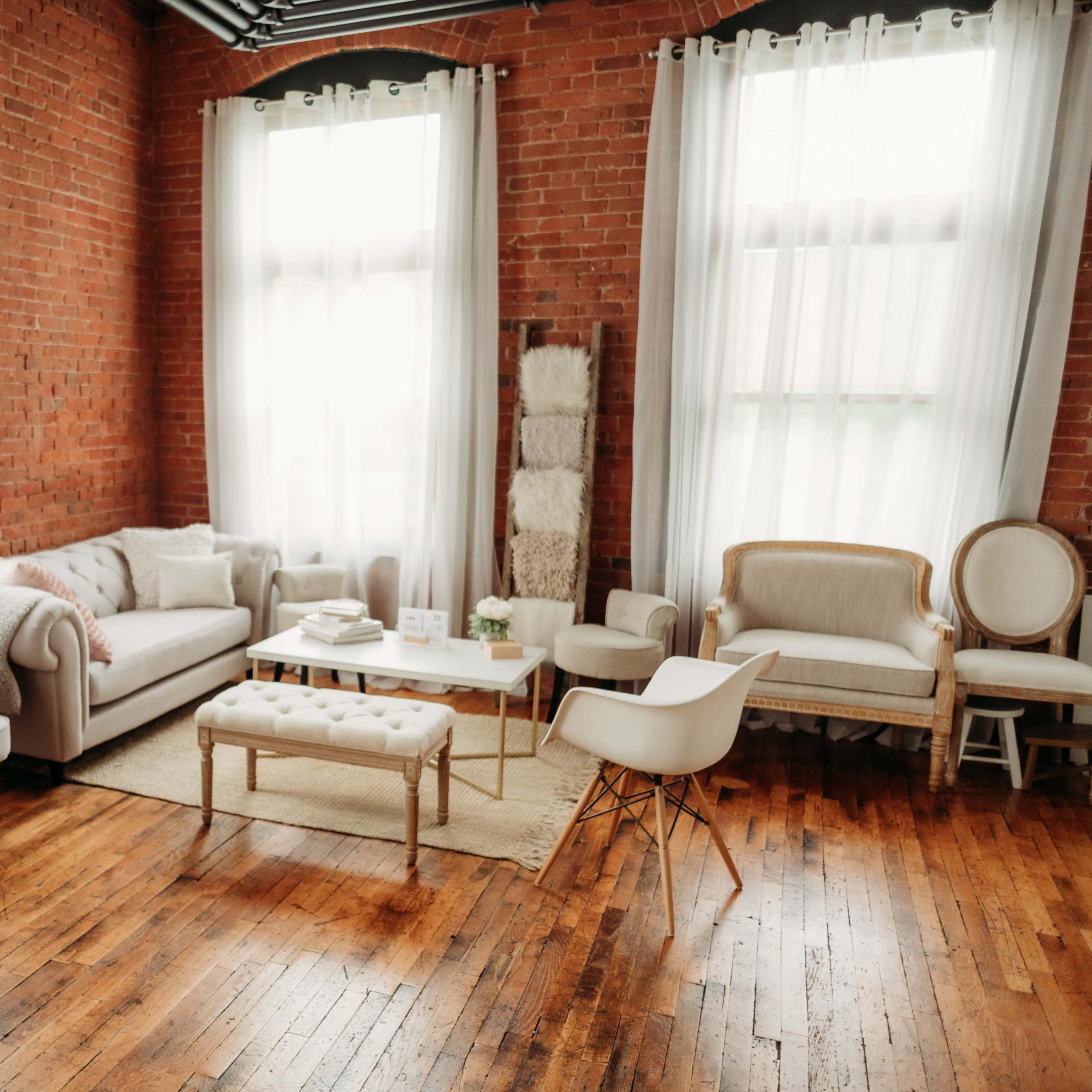 A spacious living room with exposed brick walls, large windows, and a combination of modern and vintage furniture arranged around a central coffee table.