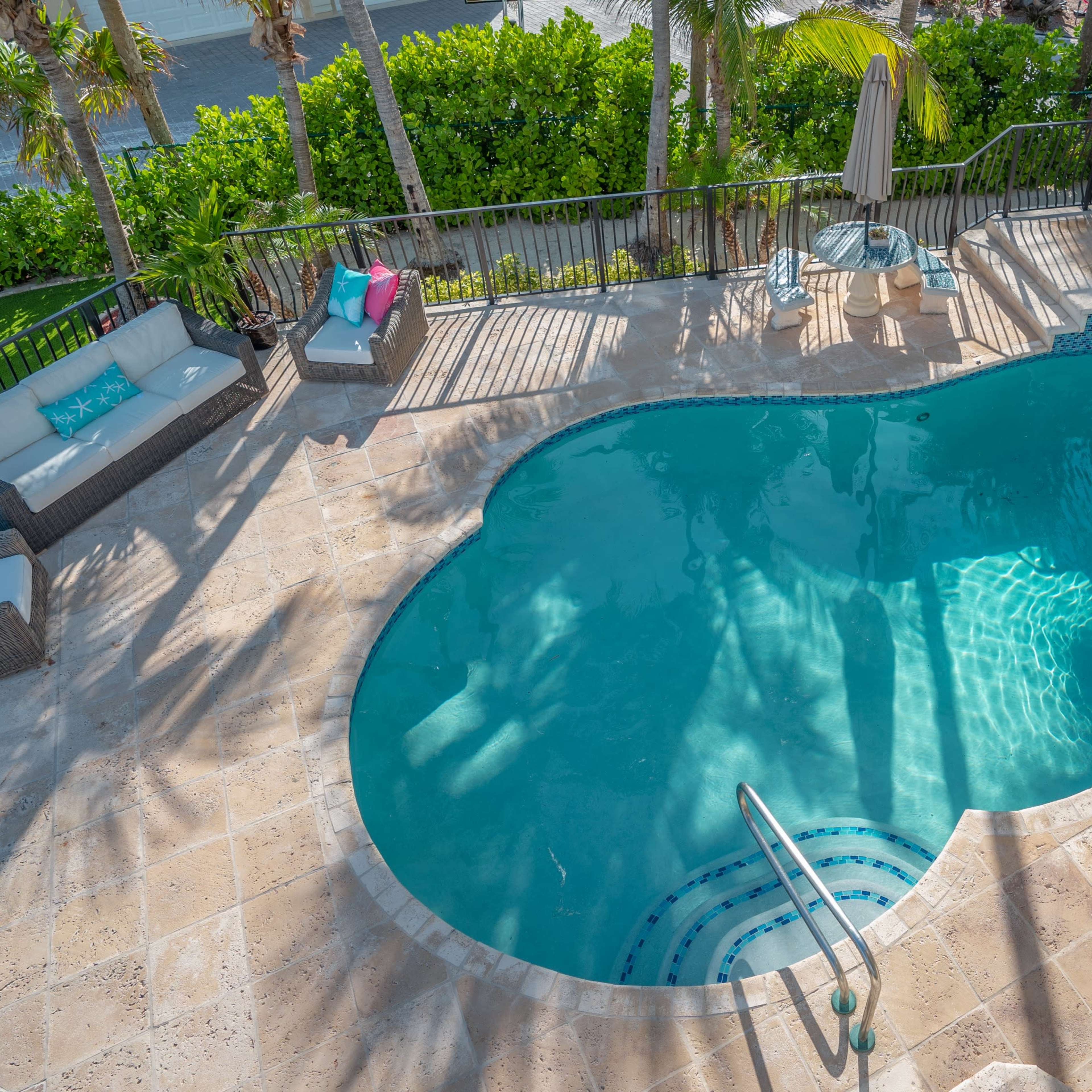 A turquoise swimming pool with a spa is surrounded by patio furniture and palm trees.