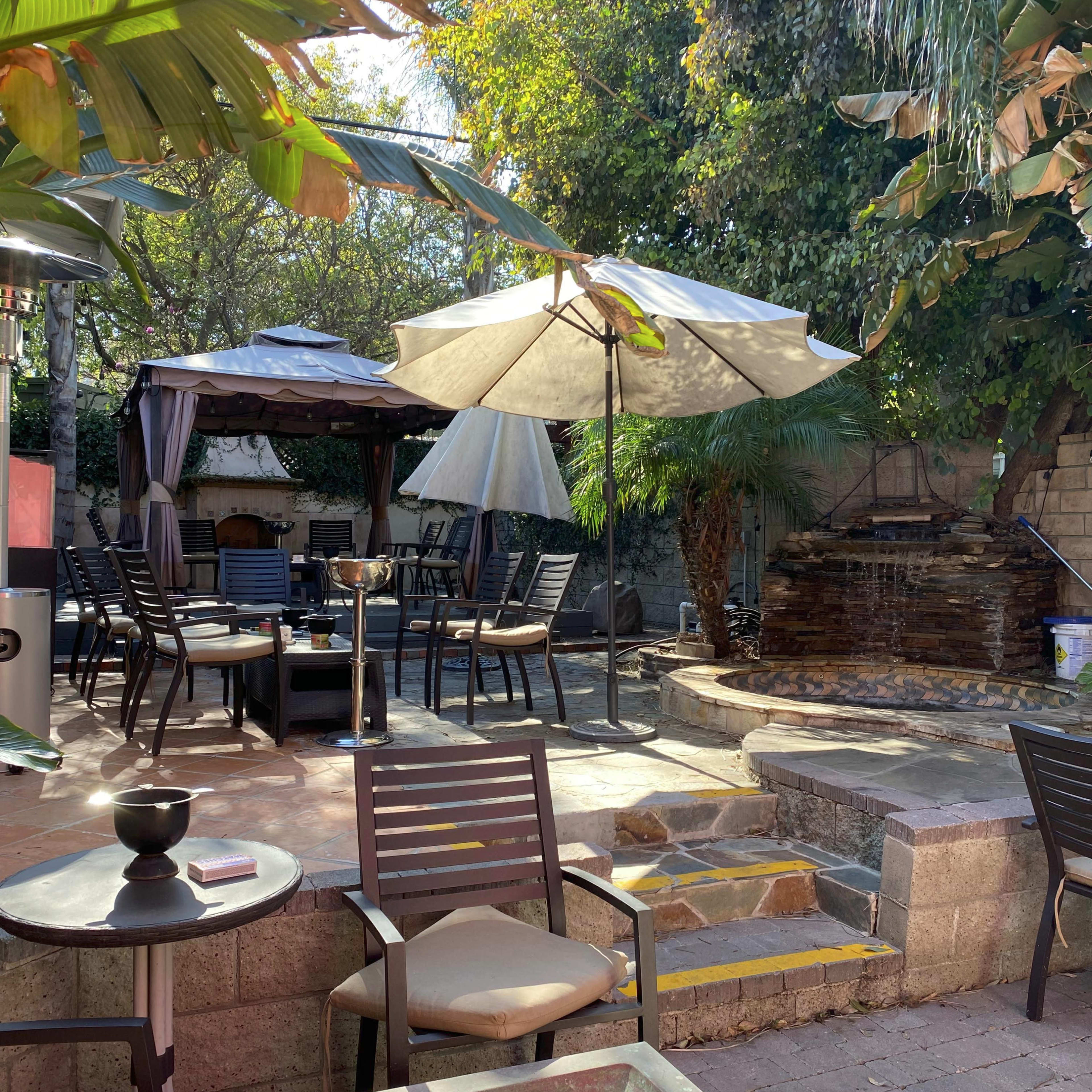 An outdoor patio area with seating, tables, a gazebo, and a water feature surrounded by greenery.