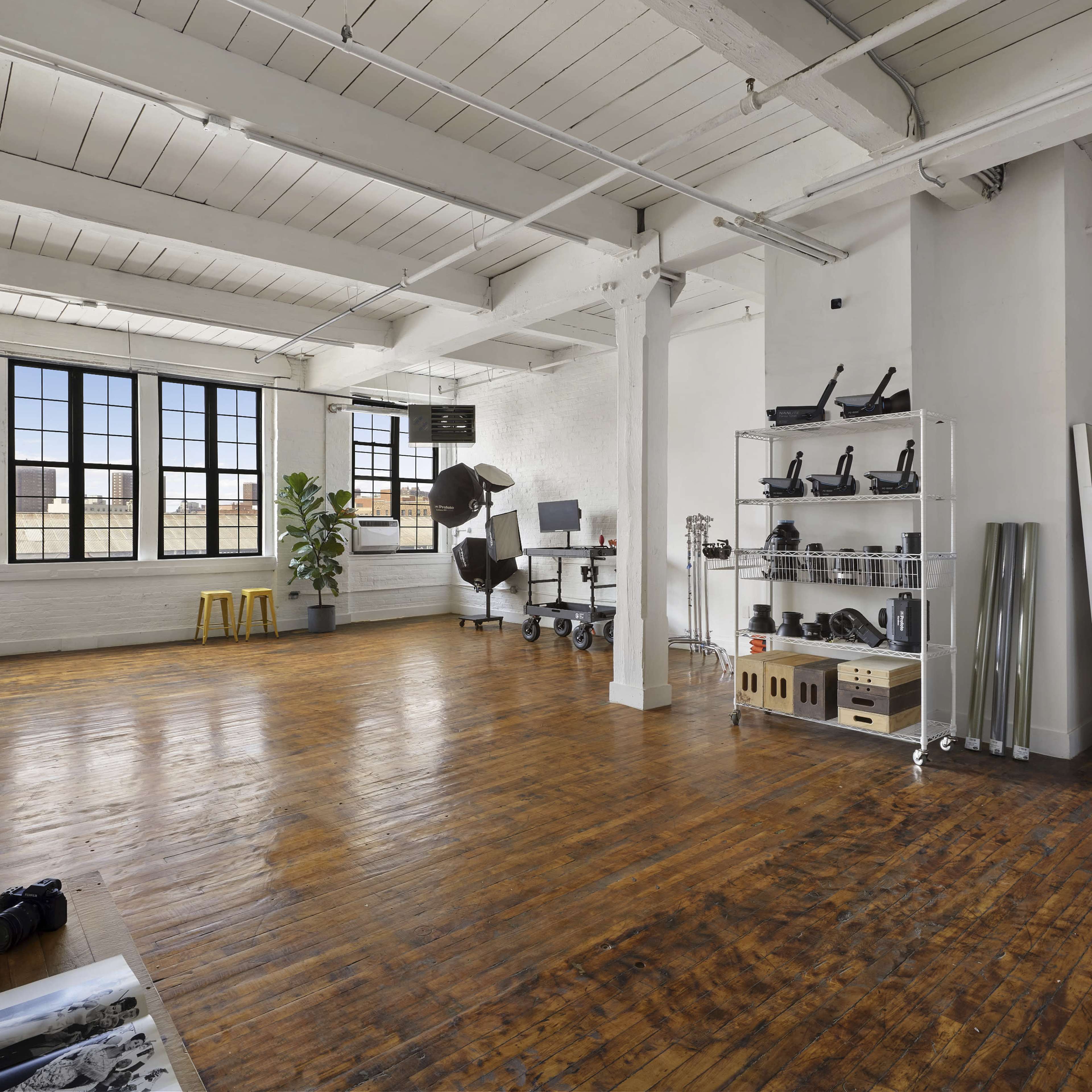 A spacious, well-lit studio with wooden floors, large windows, a variety of photography equipment, and minimalist furnishings.