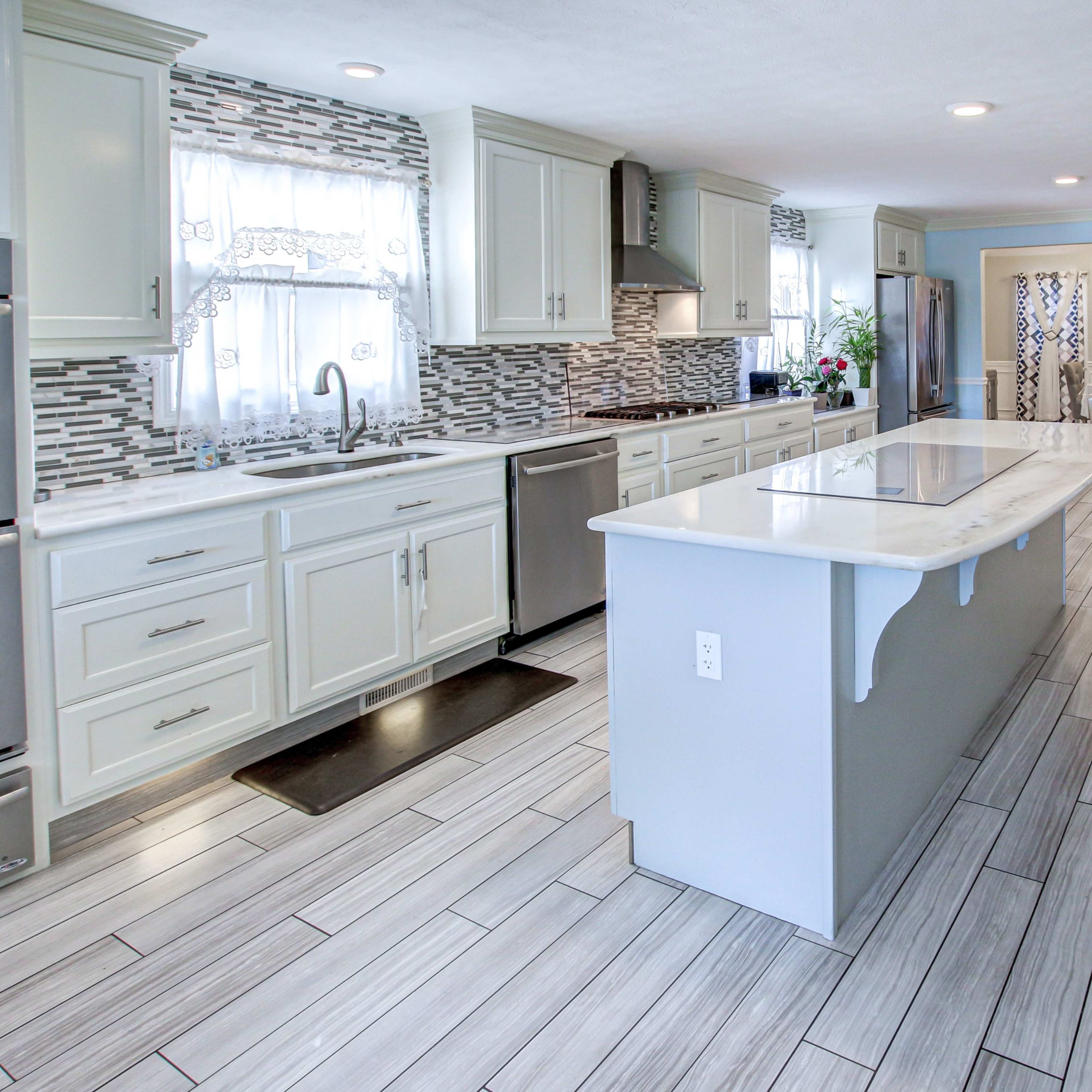 A modern kitchen with white cabinetry, a large island, and tiled flooring, leading to a dining area with a table and chairs.