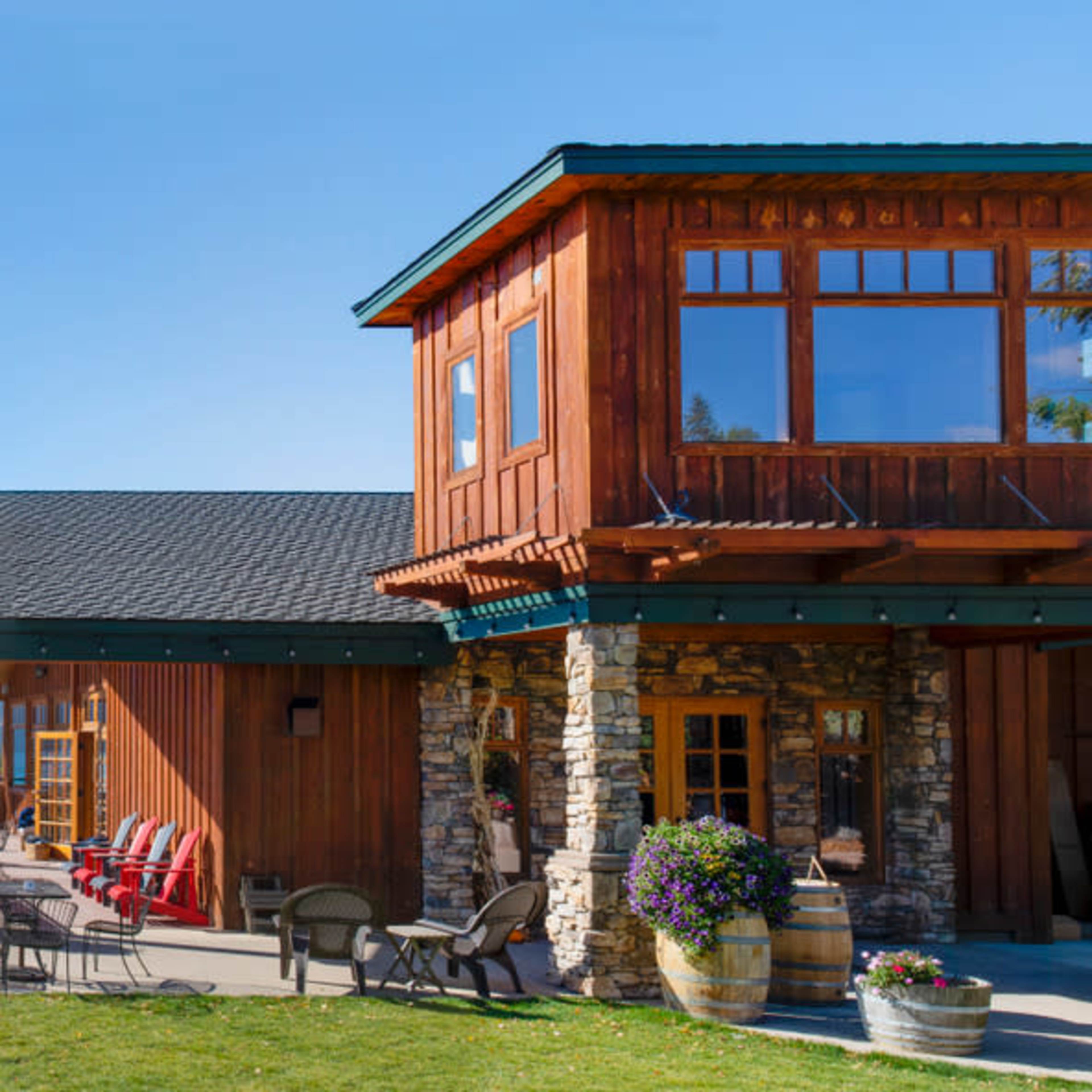 A wooden lodge with large windows overlooks a lake surrounded by mountains, with patio seating in the foreground.