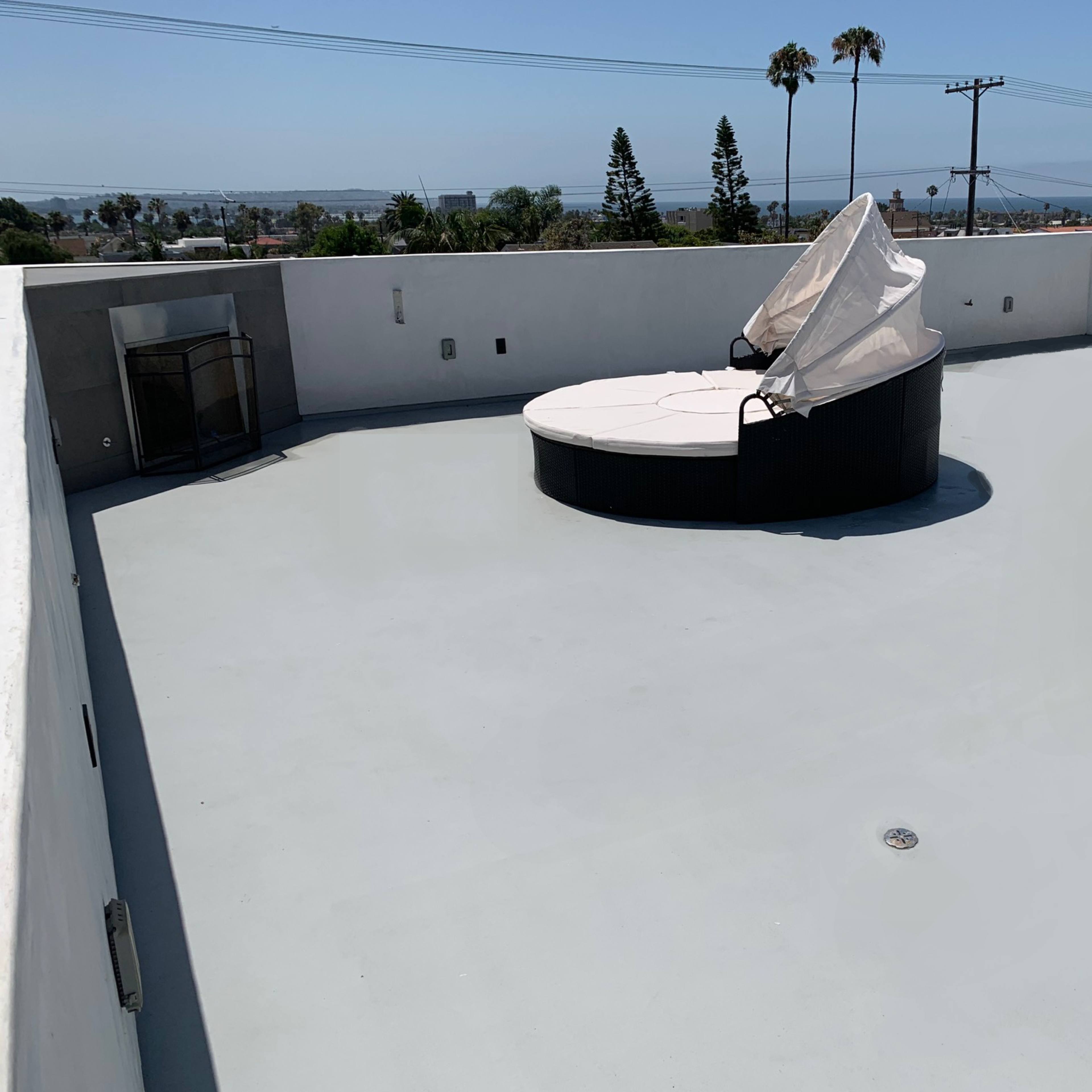 A flat rooftop features a circular seating area with a large cushion, surrounded by palm trees and a clear view of the ocean in the background.