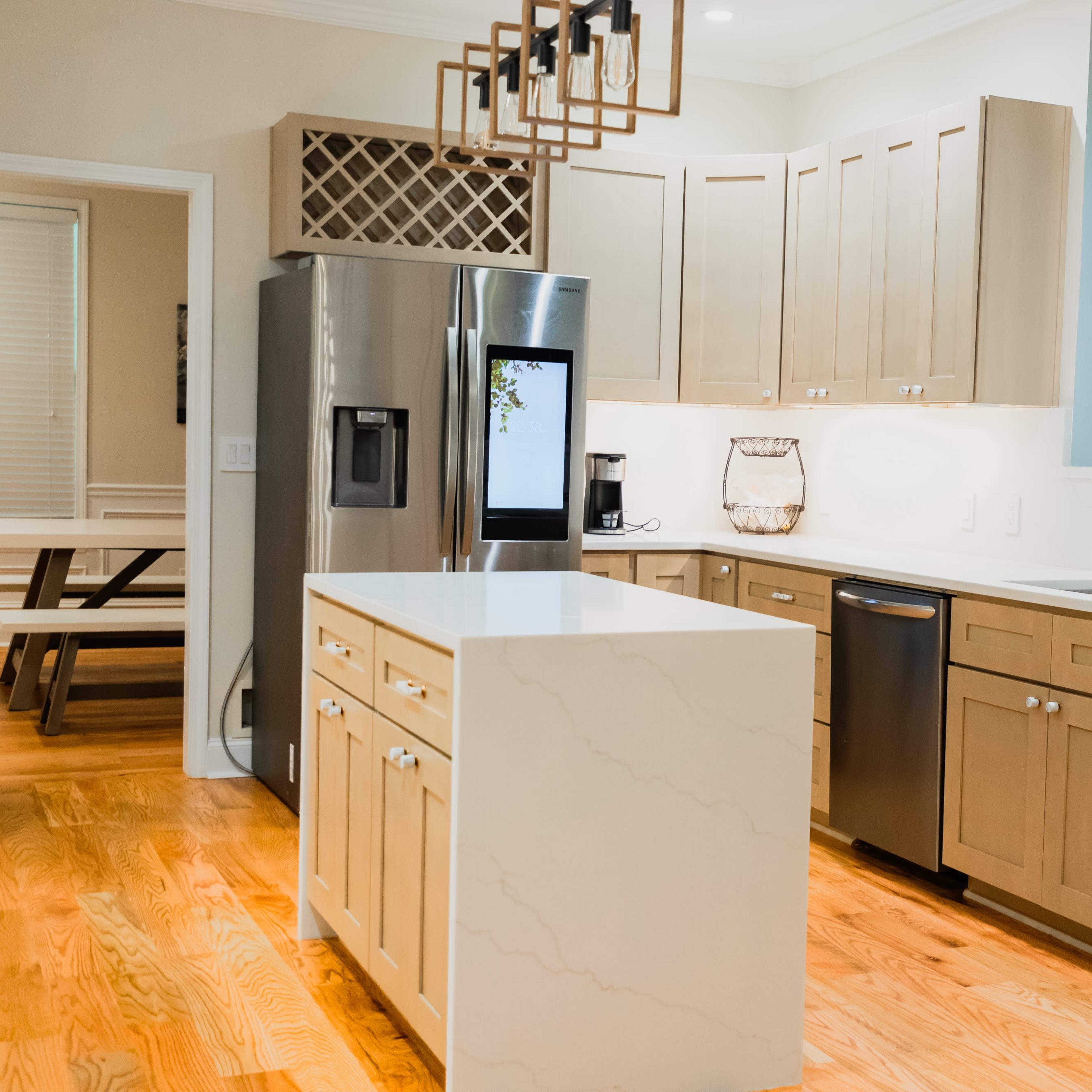 A modern kitchen with light wood cabinets, stainless steel appliances, a central island with a white countertop, and hardwood flooring.