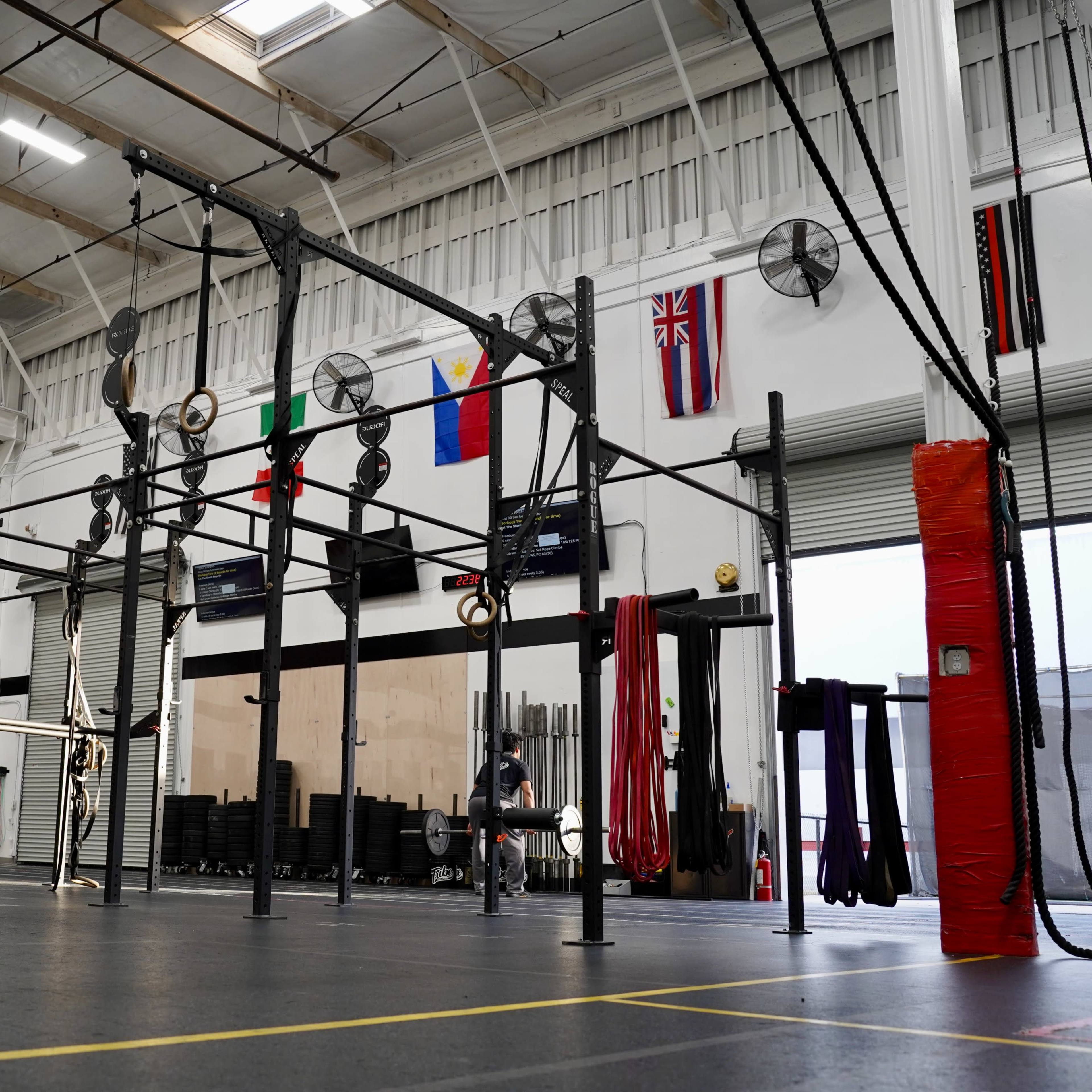 A spacious gym interior with various exercise equipment, including pull-up bars and weights, under a high ceiling adorned with flags from different countries.