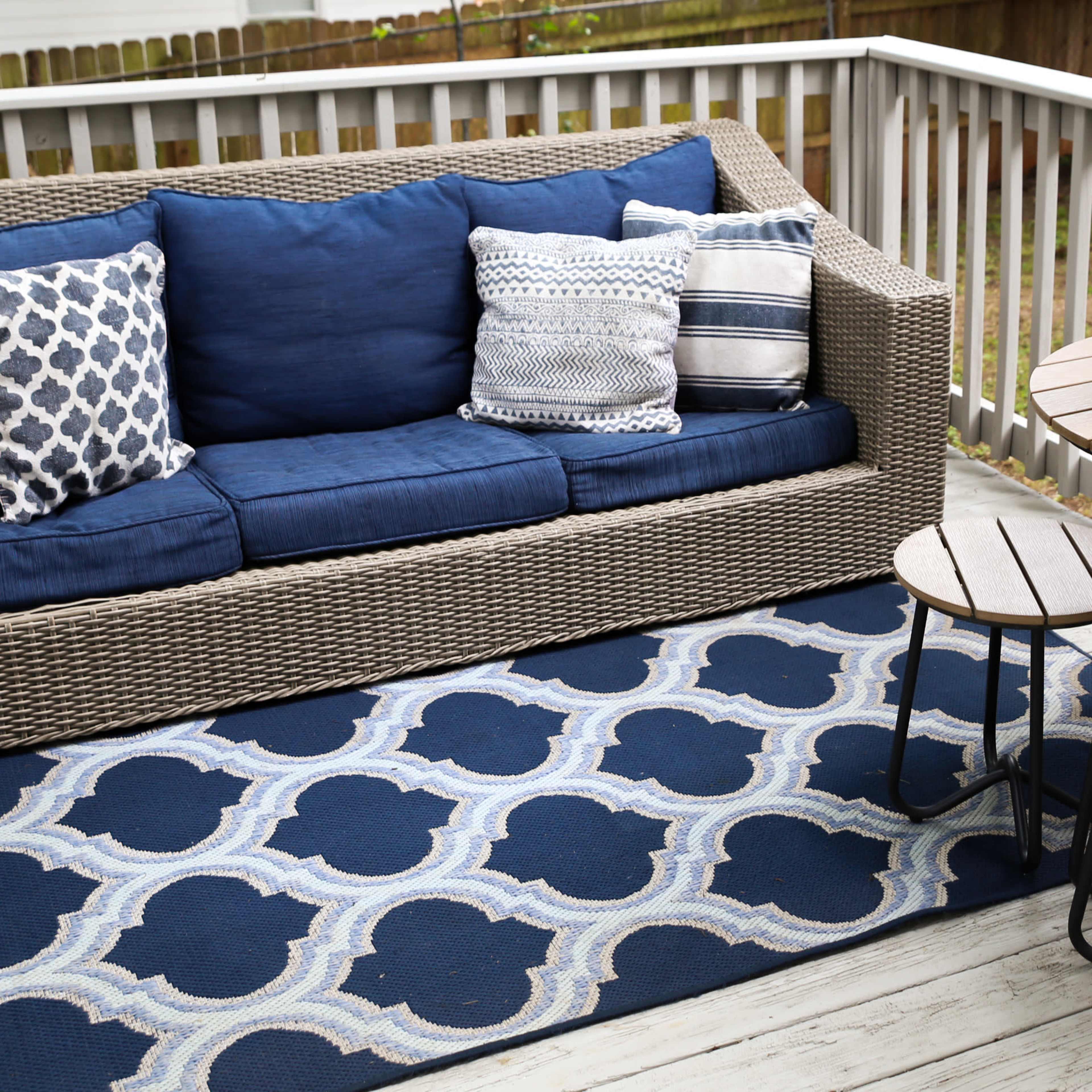A wicker sofa with blue cushions and decorative pillows sits on a deck alongside a small round table and four chairs, with yellow tulips in a vase.