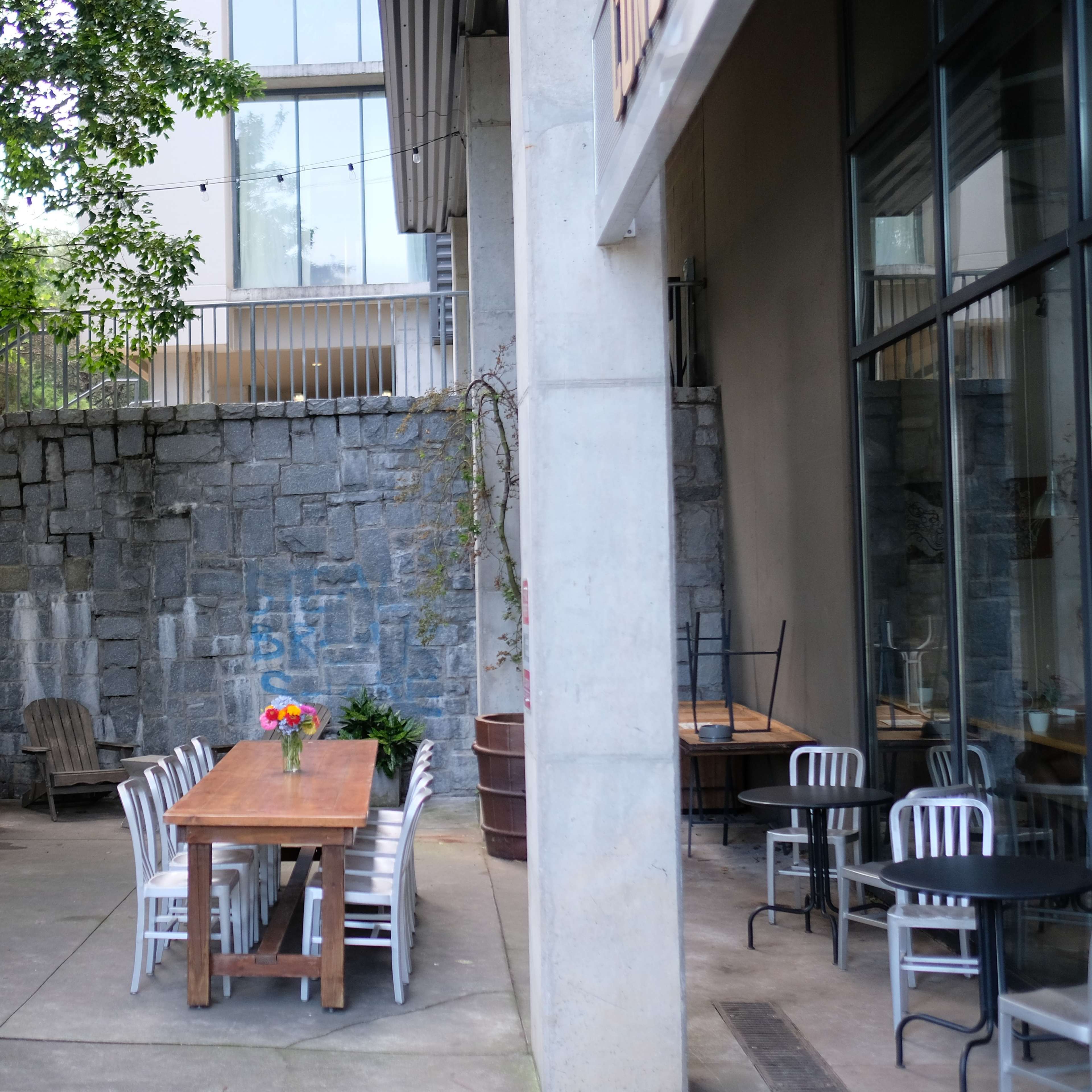 An outdoor dining area features a long wooden table with flowers surrounded by benches and metal chairs against a stone wall.