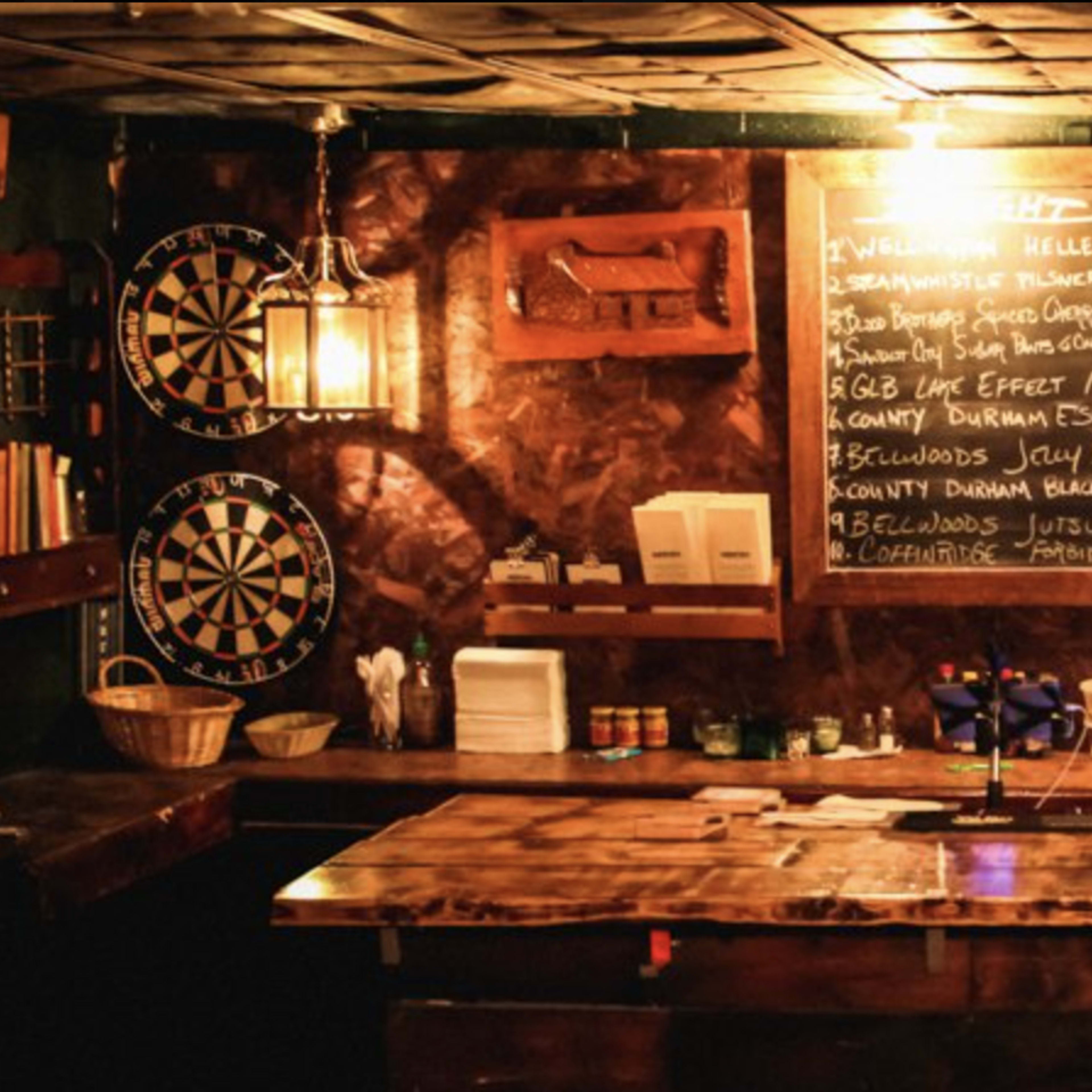 A wooden bar is set against a dark wall adorned with a chalkboard menu and shelves of various liquor bottles, while dartboards are visible on either side.
