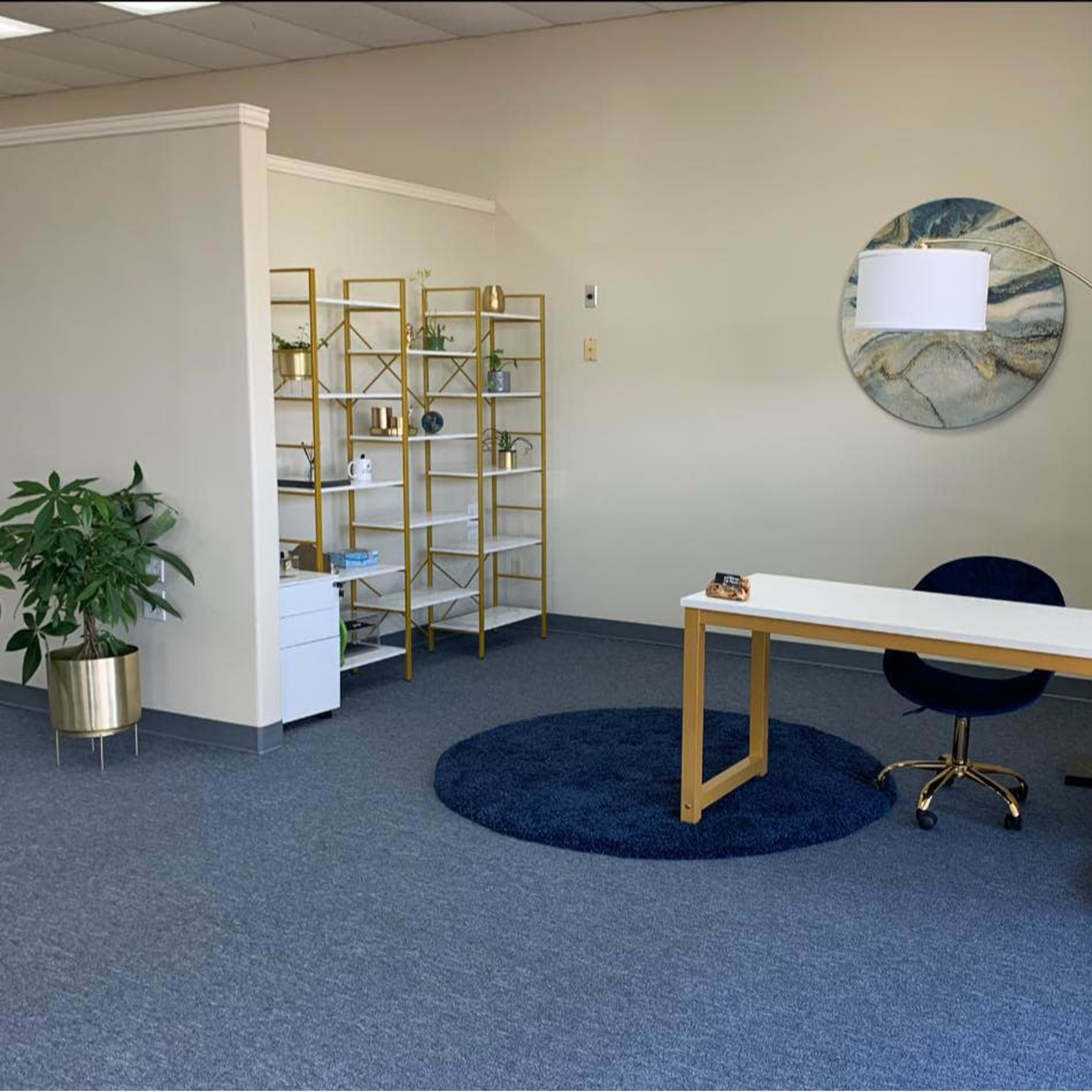 A modern office space featuring a white desk with a dark blue chair, a round rug, a bookshelf with decorative items, and a floor lamp.