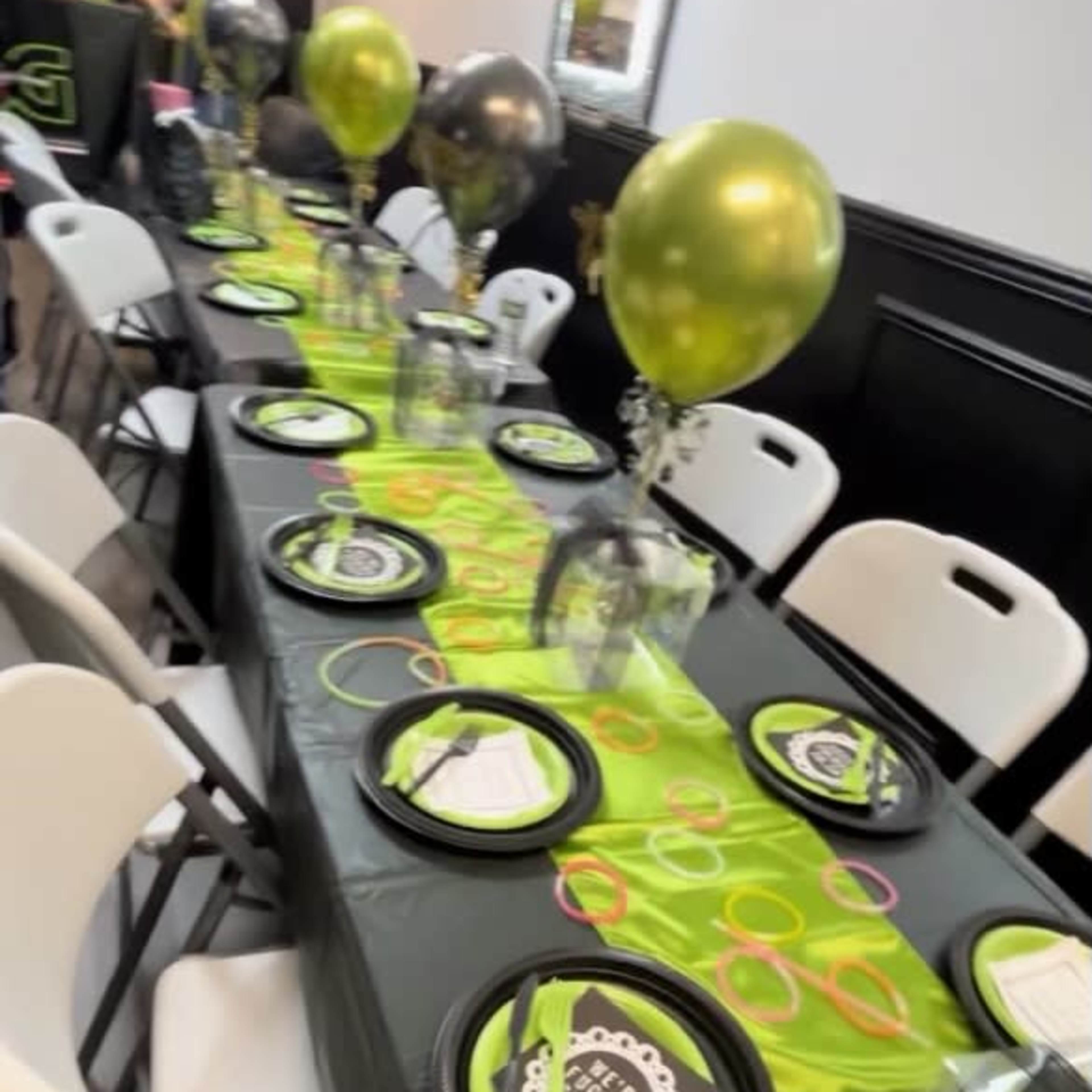 A long table is set for a party, adorned with green and black tableware and decorated with balloons.