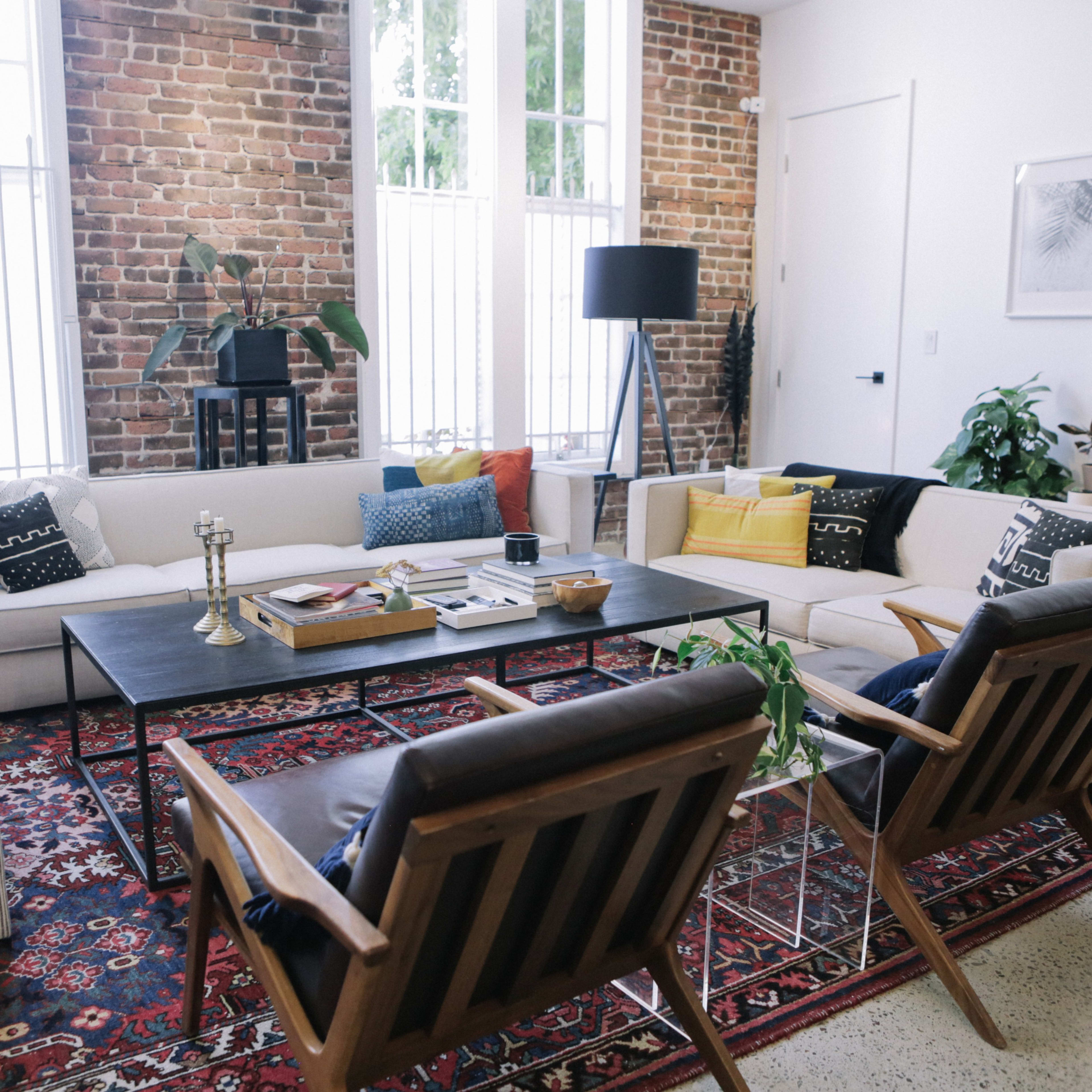 A modern living room features a mix of seating options, including a sectional sofa and two wooden chairs, arranged around a central coffee table on a patterned rug against a backdrop of exposed brick walls and large windows.