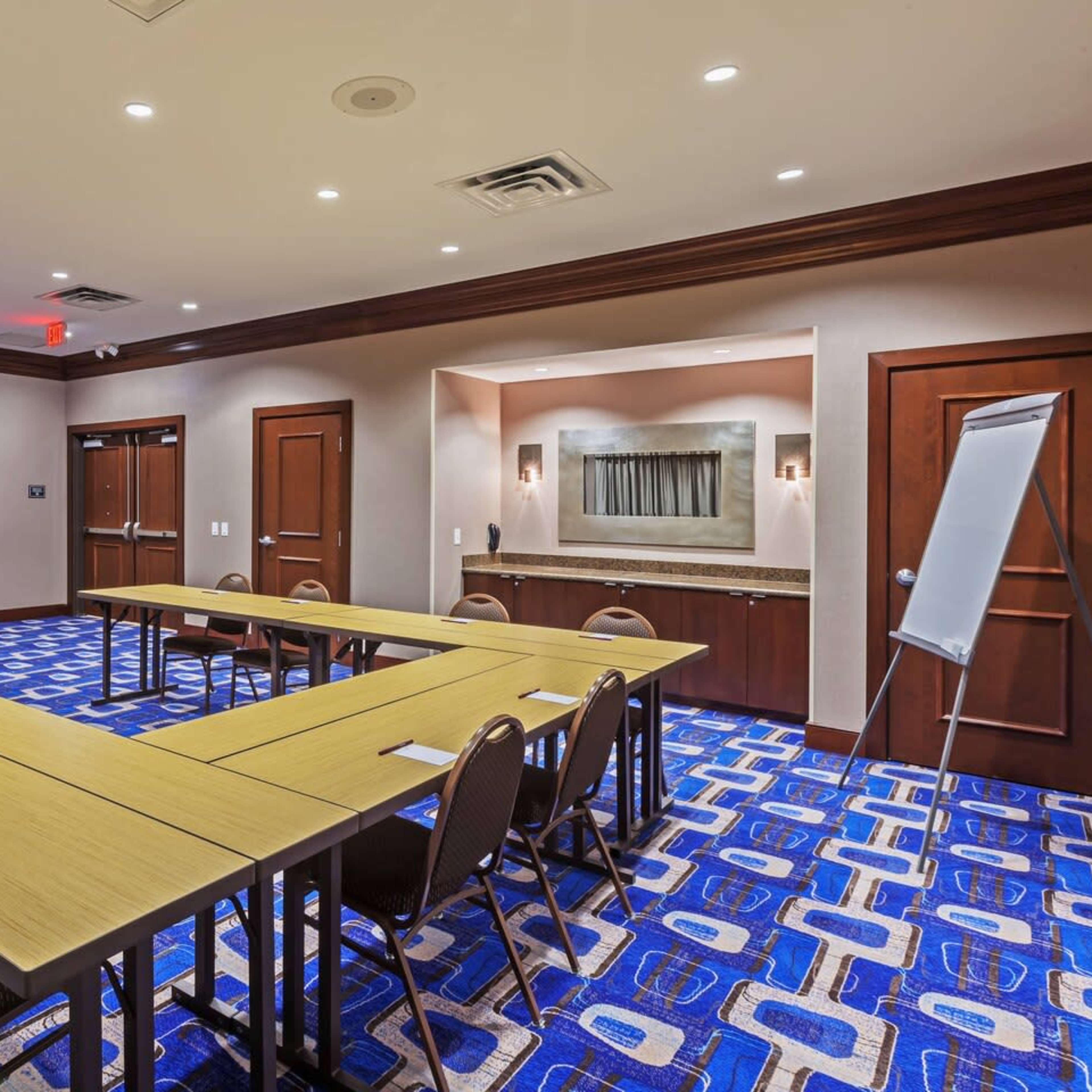 A conference room features a U-shaped seating arrangement with tables and chairs, illuminated by overhead lighting and decorated with a patterned carpet.